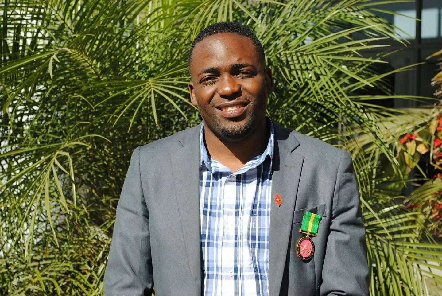 A man in a gray suit with a checked shirt standing outdoors in front of green tropical plants, smiling at the camera with medals pinned on his jacket.