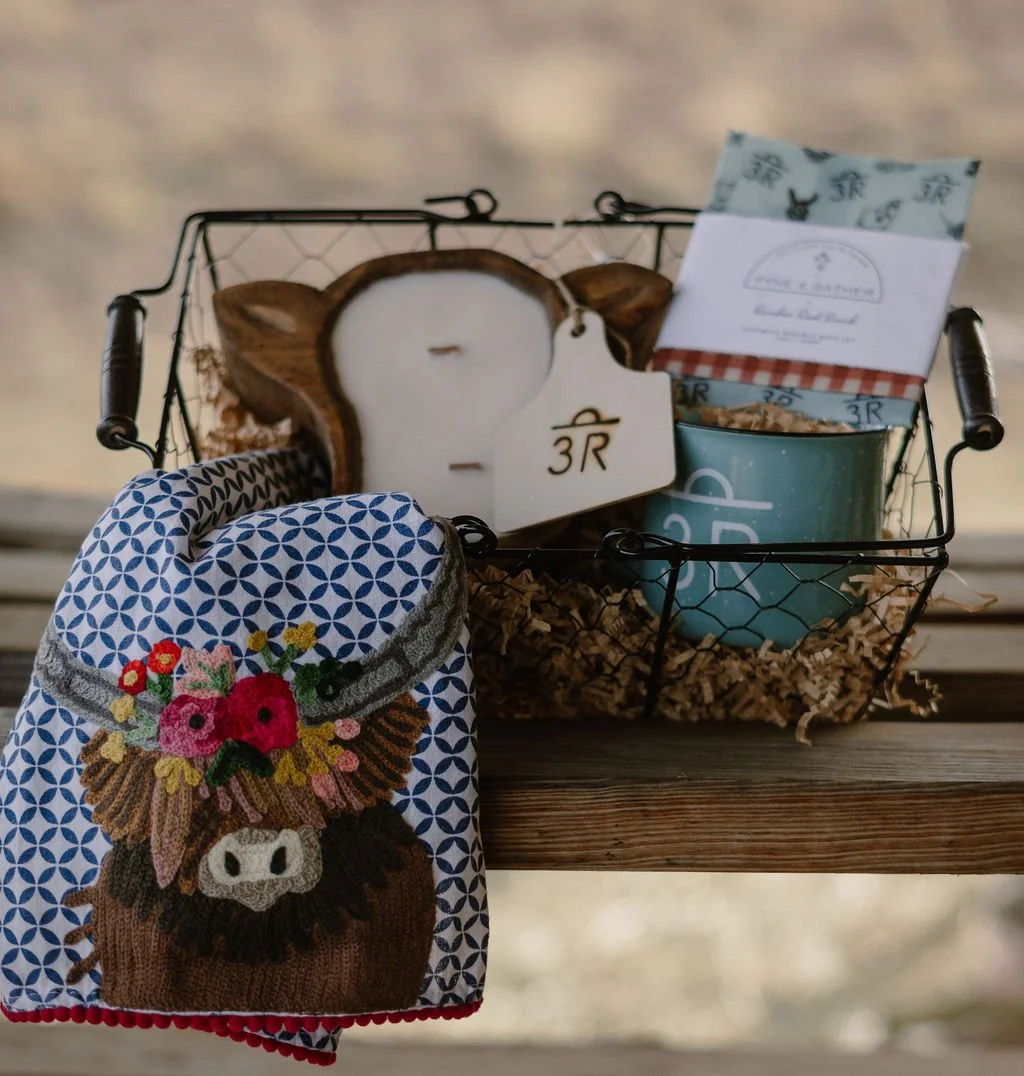 A wire basket holding candles, tea, and snacks, with a decorative oven mitt featuring a cow and floral embroidery placed on a wooden surface.