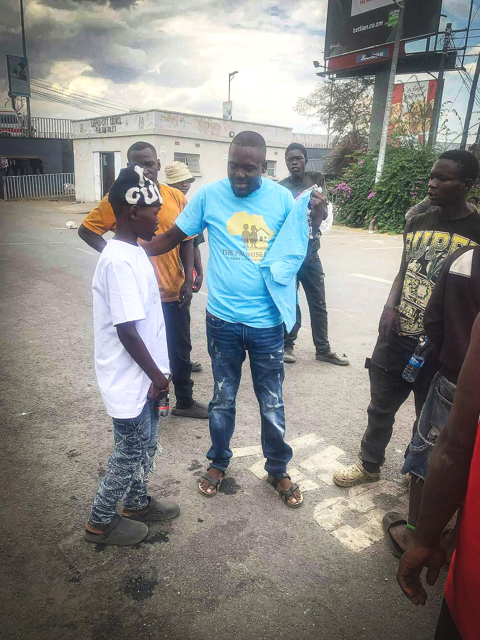 A group of six men standing on a street in daytime. One man in a blue T-shirt is handing an item to a boy in a white T-shirt. The men are casually dressed, and there are buildings and billboards in the background.