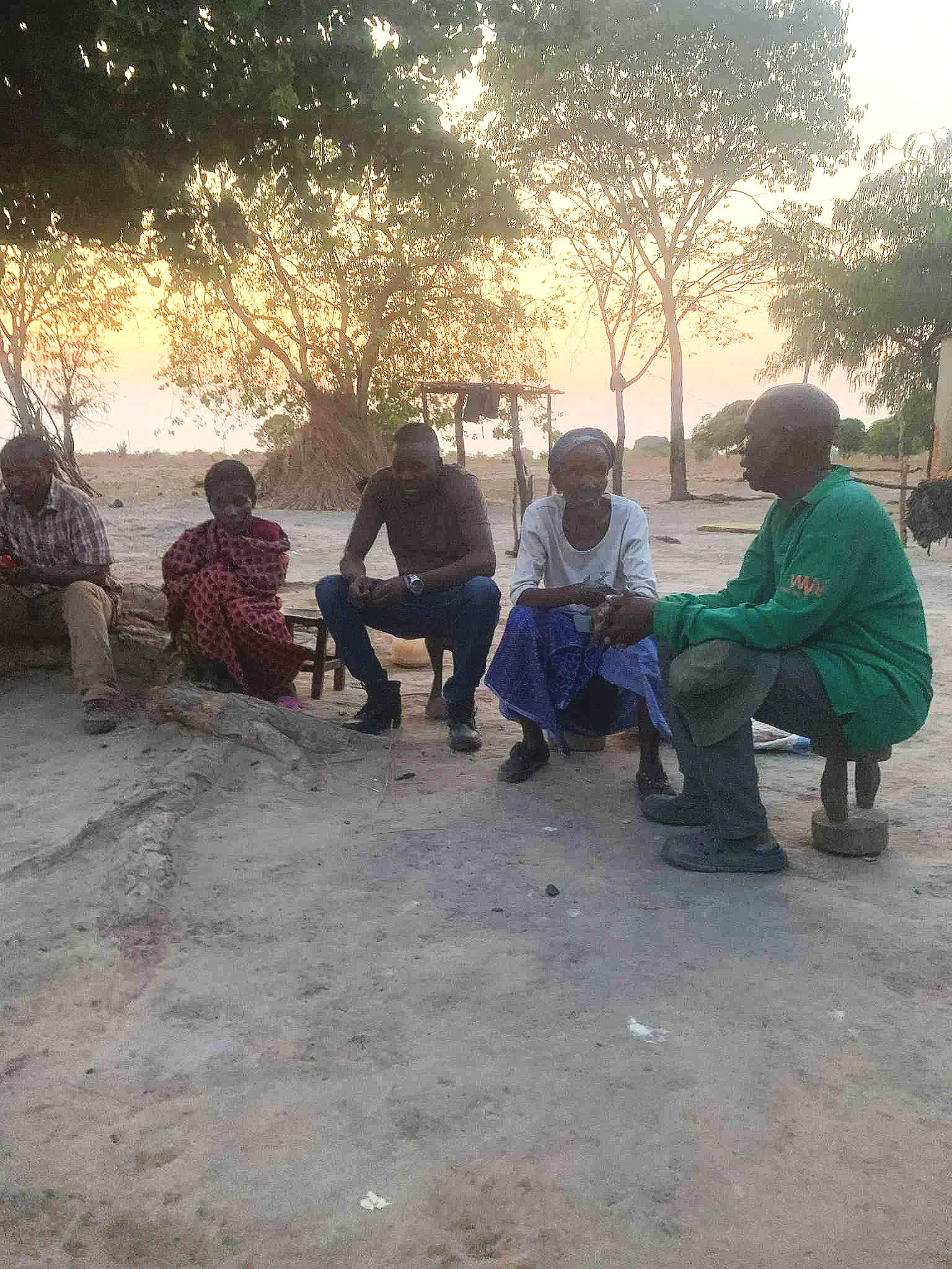 Group of five people sitting outdoors on benches and rocks, shaded by trees, during sunset in a rural setting.