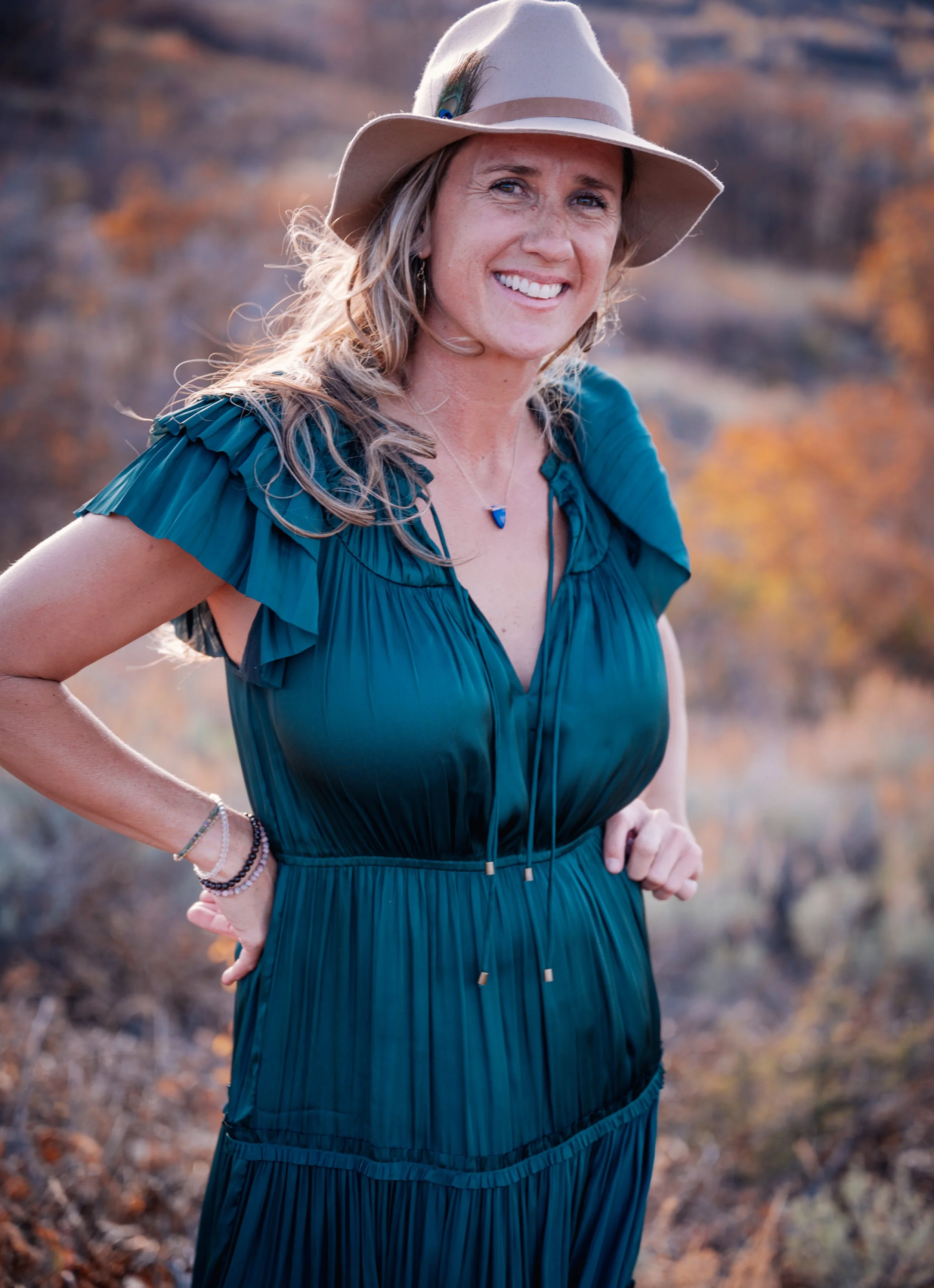 A woman outdoors wearing a teal dress and a beige wide-brimmed hat, smiling, with autumn trees in the background.