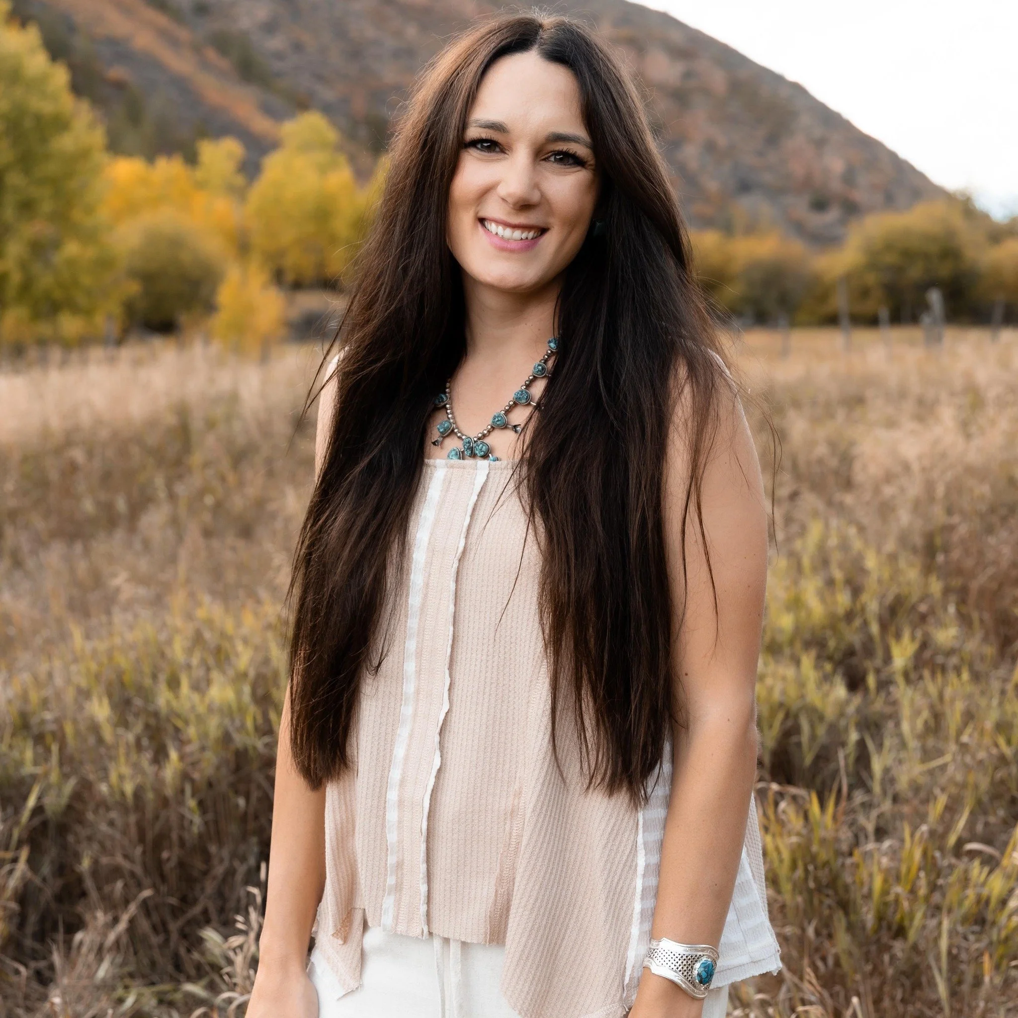 A woman with long dark hair smiling outdoors during fall, wearing a sleeveless beige top, turquoise jewelry, and a silver bracelet, with a mountain and autumn trees in the background.