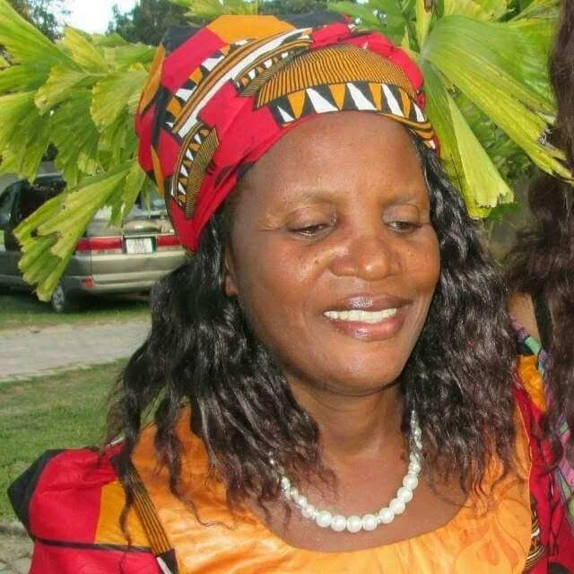 A woman wearing a colorful headwrap and necklace, standing outdoors with green leaves and cars in the background, smiling slightly.