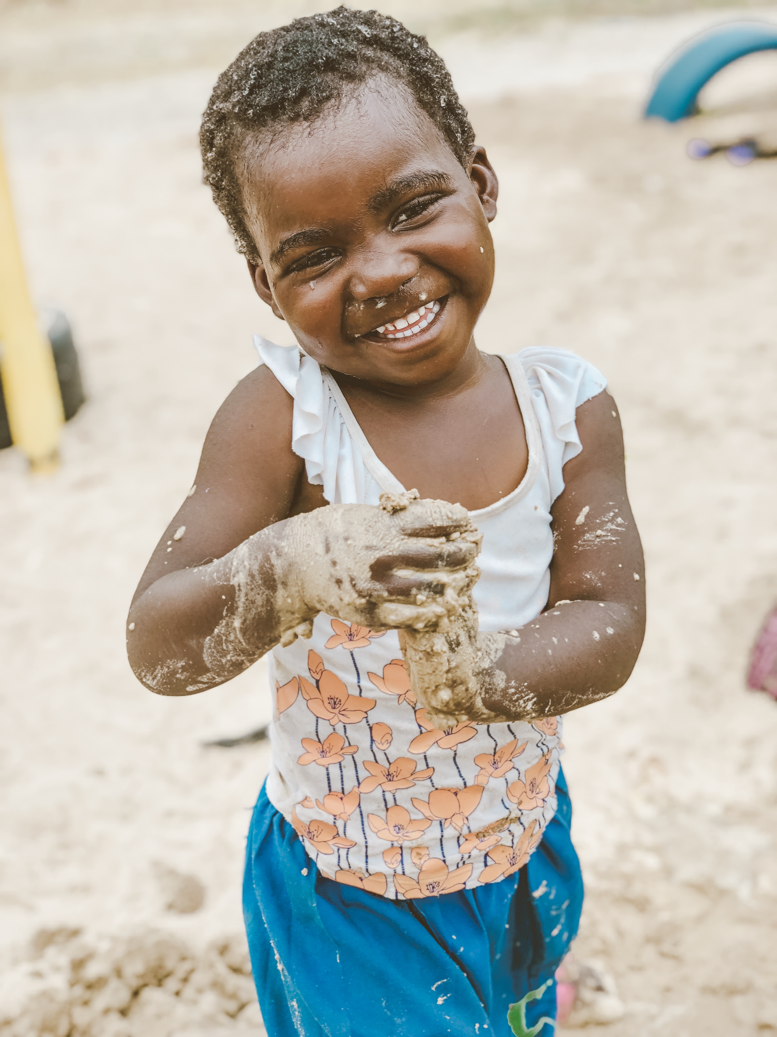 A smiling young girl with dark curly hair, wearing a white and orange floral top and blue shorts, playing in sand and holding a clump of wet sand in her hands.