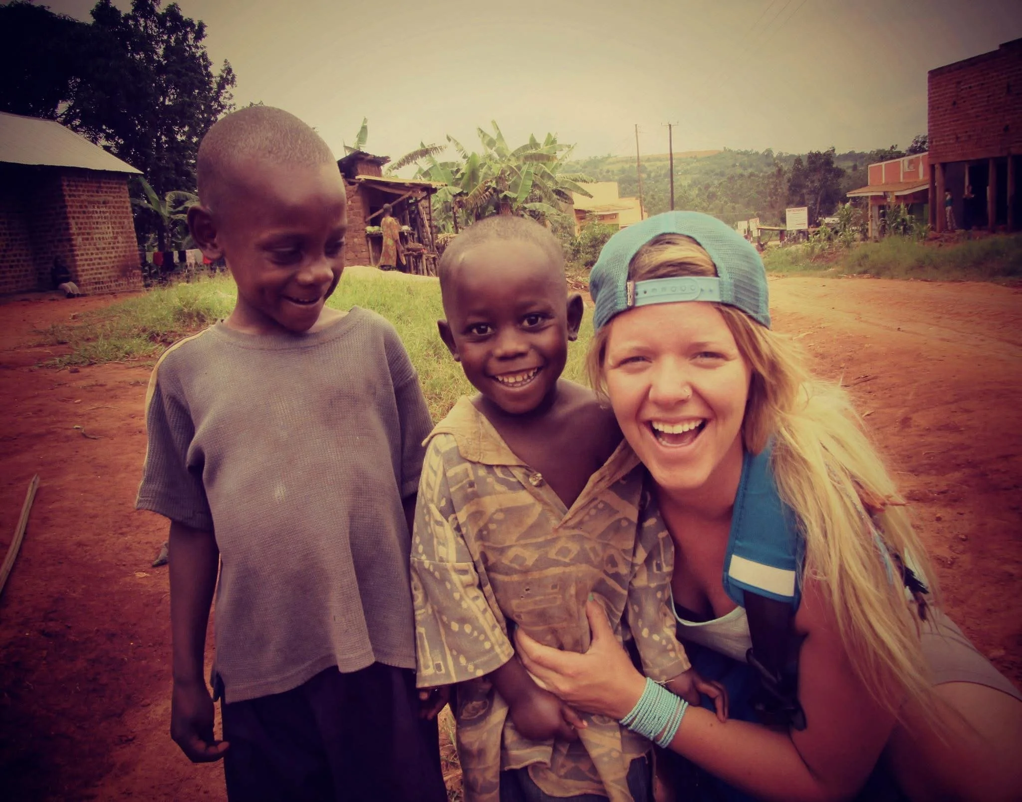 A woman with long blonde hair wearing a blue backward cap and a blue backpack smiling and hugging a young boy with a shaved head and patterned shirt, standing on a dirt road with two other children in a rural area with houses and banana plants in the background.