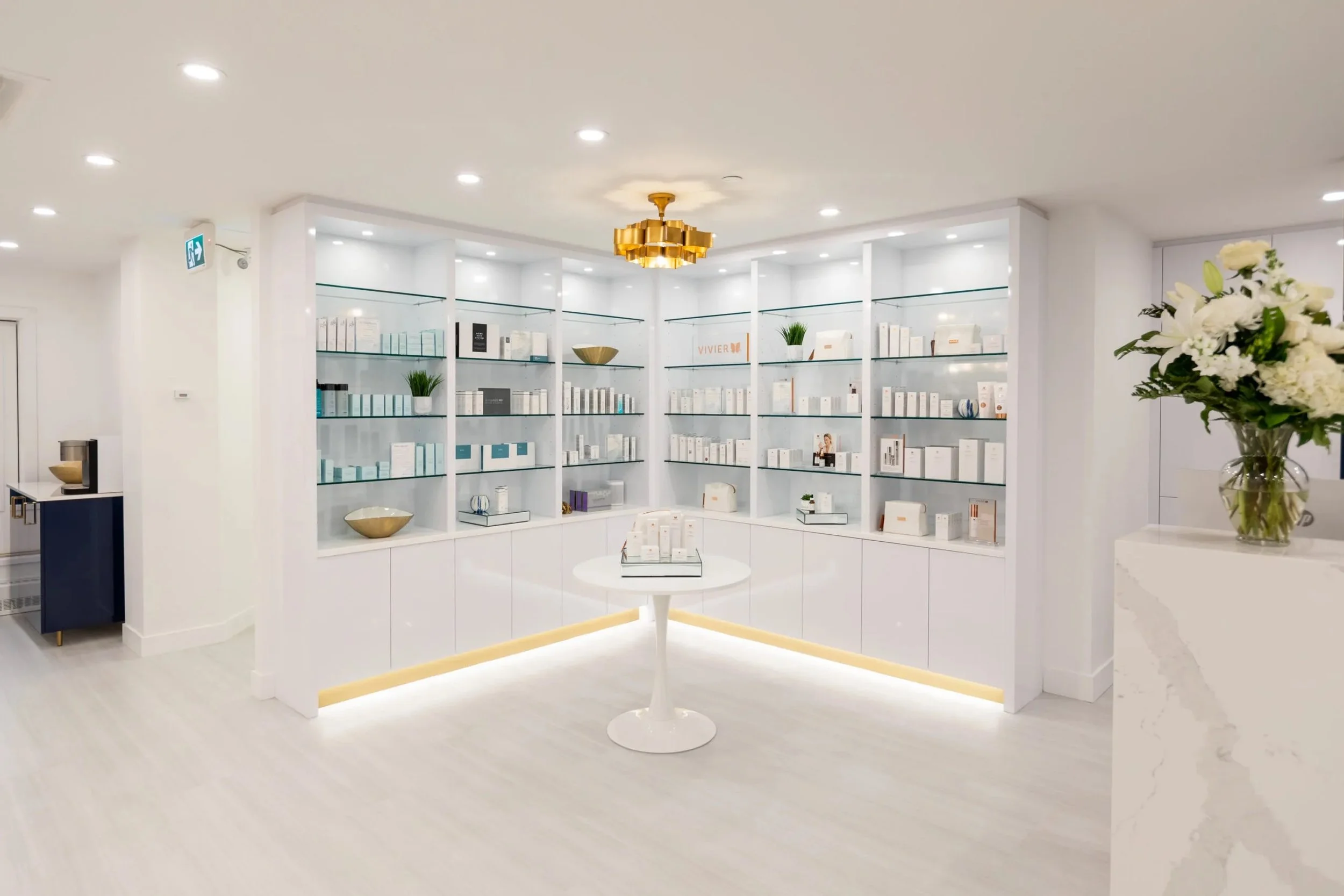 An interior space with white shelves filled with skincare or cosmetic products, a small round table with product displays, and a vase of white flowers on a white counter.