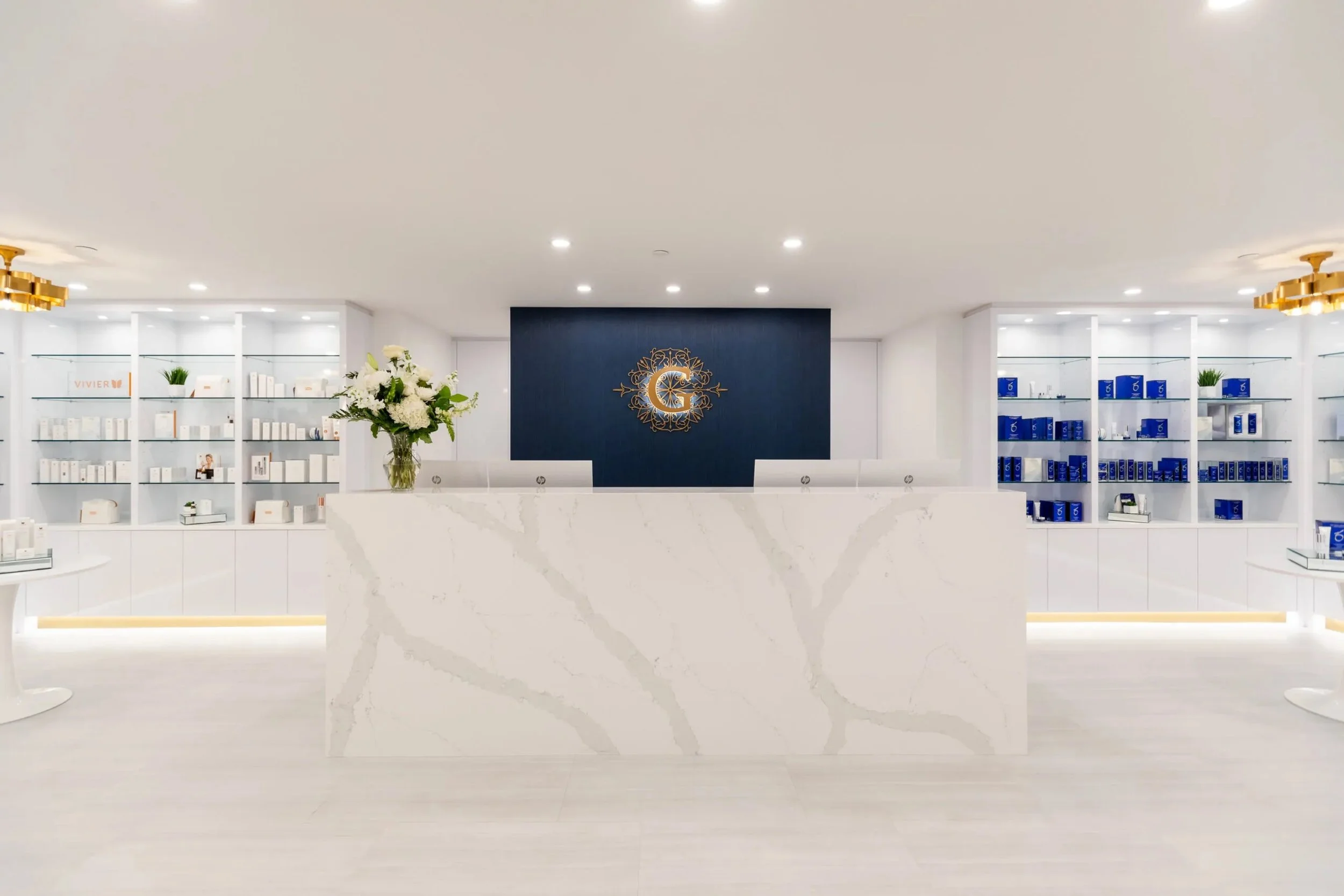 Modern reception area with a white marble front desk and navy blue accent wall featuring a gold decorative clock. White shelves with skincare products and blue boxes flank the space, decorated with small potted plants, and a glass vase with white flo