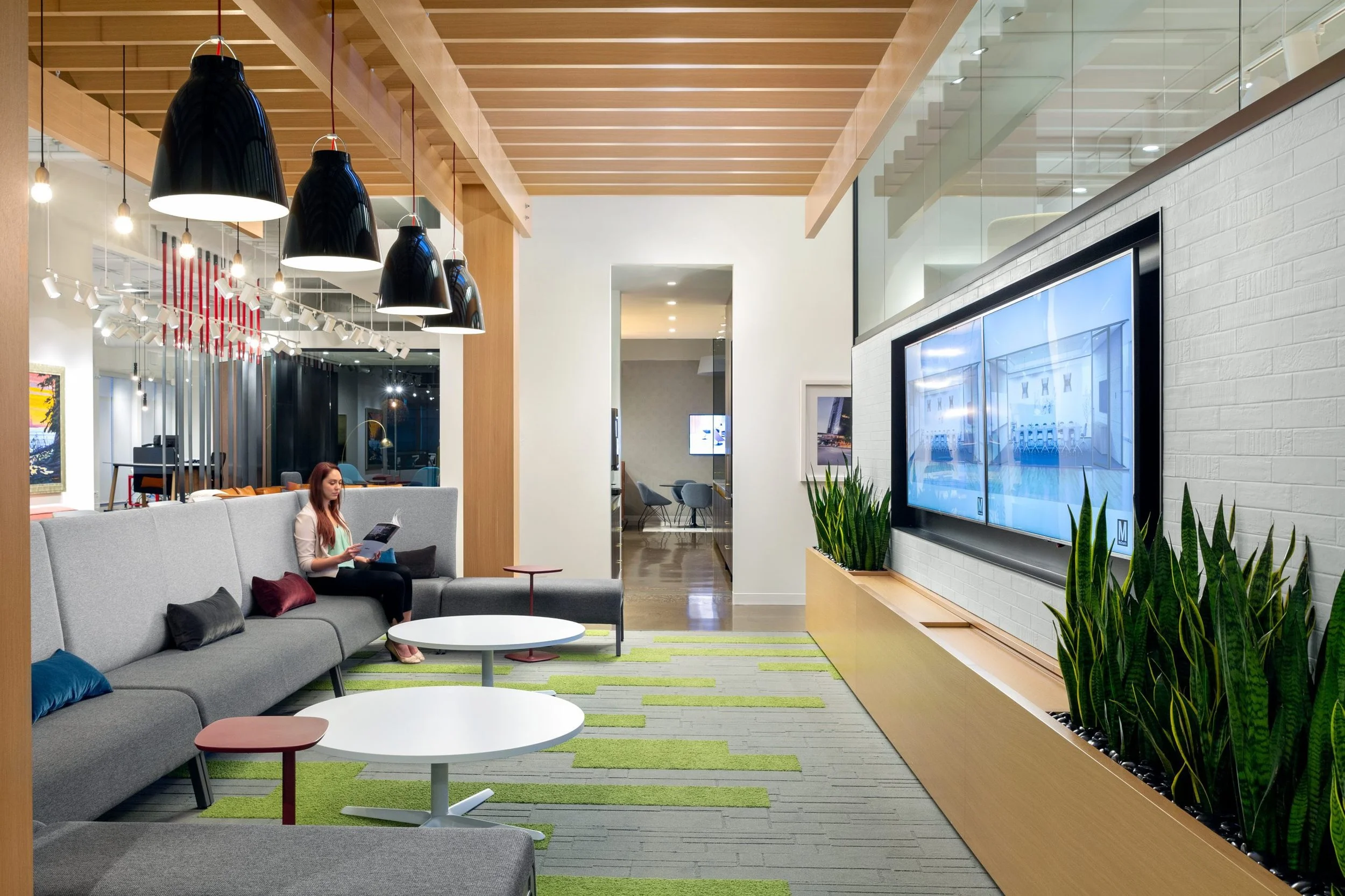 Modern hotel lobby with a gray sofa, a woman reading a magazine, black pendant lights, and a large television screen on a white brick wall surrounded by green plants.