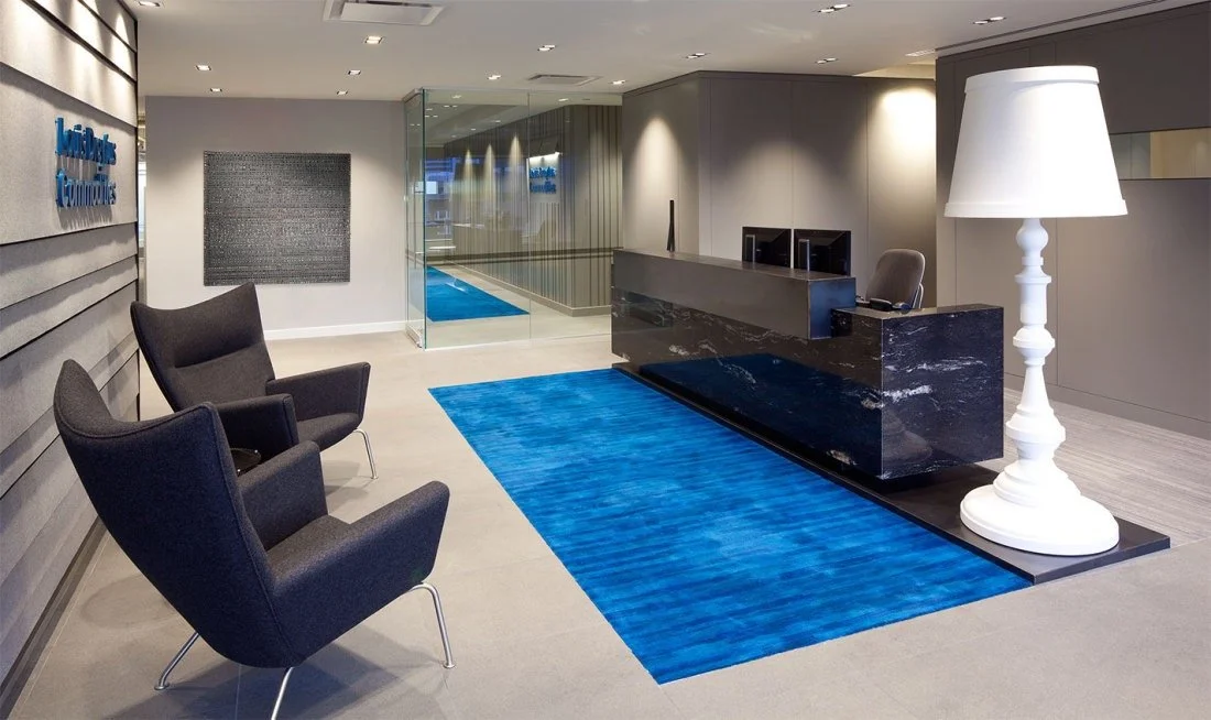 Modern hotel lobby with a reception desk, seating area with black armchairs, blue rug, large white floor lamp, and a glass wall with reflections.