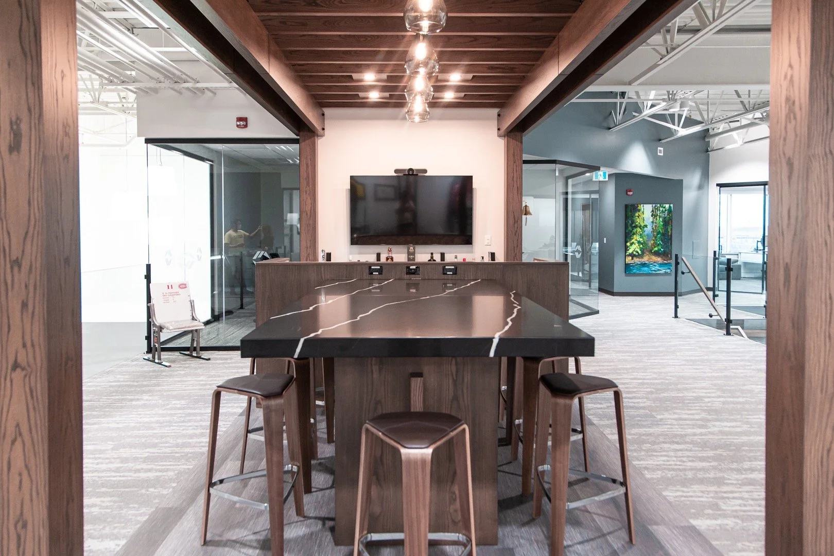 Modern interior of a conference room with a black marble table, wooden chairs, a wall-mounted flat-screen TV, and a gallery wall with artwork, with glass walls and wooden beams.