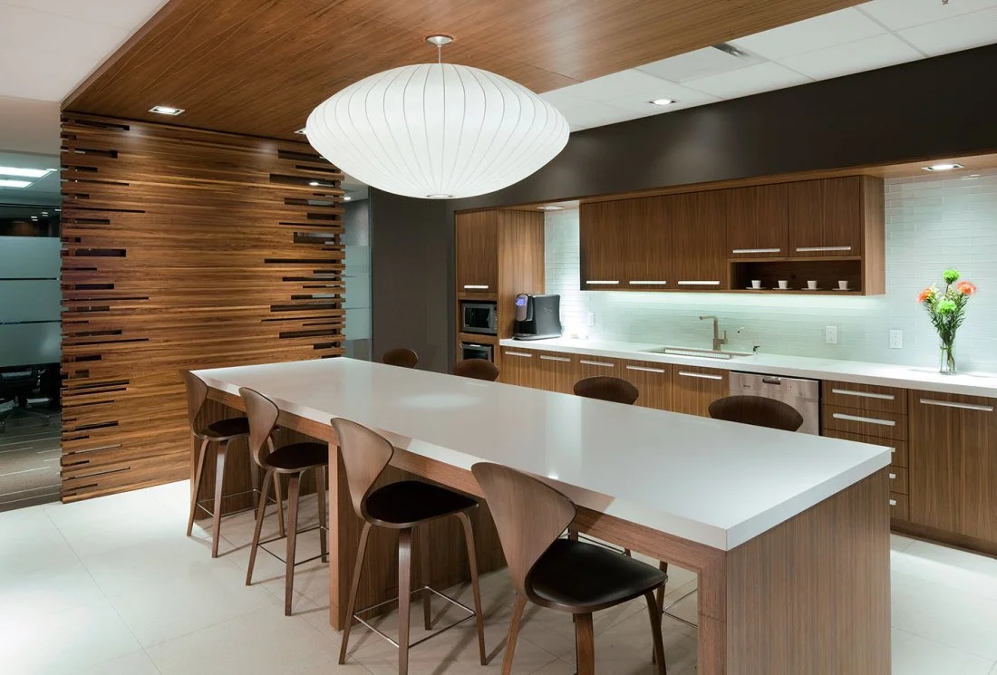 Modern kitchen with wooden cabinets, a large white island counter, and a wooden slatted wall panel. There are six wooden chairs around the island. The kitchen has a hanging paper lantern light and a vase of colorful flowers on the countertop.