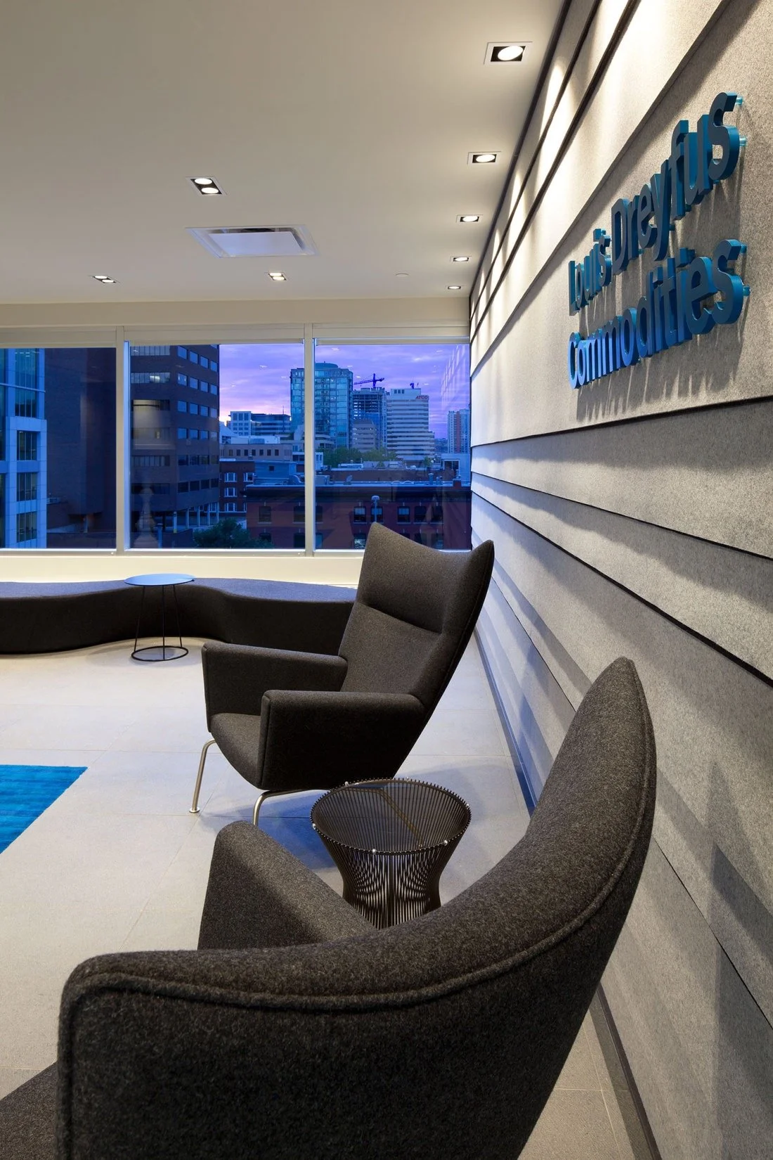 Modern office lounge with black chairs, a small side table, and large windows revealing a cityscape at dusk, with a sign on the wall reading "Luis Dreyfus Commodities."