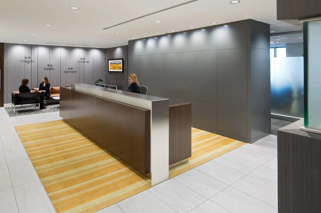 Modern office reception area with a dark wood desk, gray wall, and seated receptionist. In the background, two women are talking in a seating area with chairs and wall décor that reads 'Integrity,' 'Safety,' and 'Harmony'.