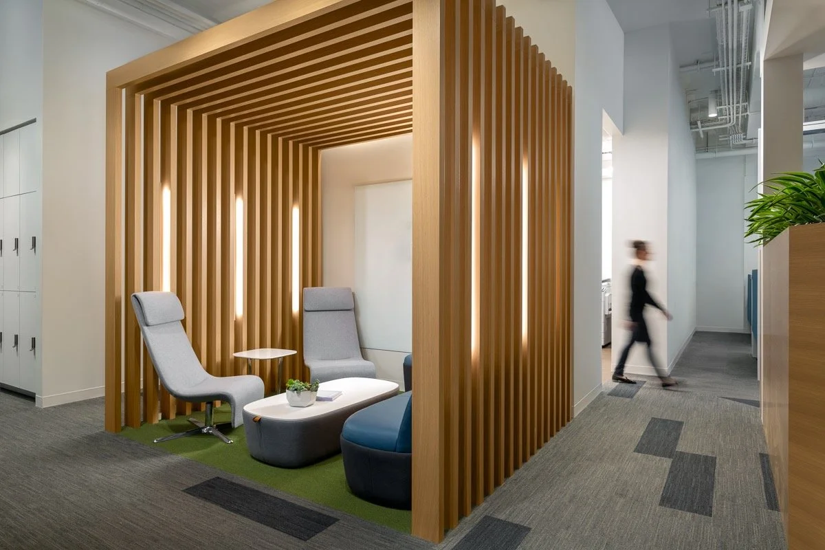 Modern office lounge area with two gray high-back chairs, a small white table, and a blue chair, enclosed by wooden slats with integrated lighting, on a green carpet inside a corporate office space.