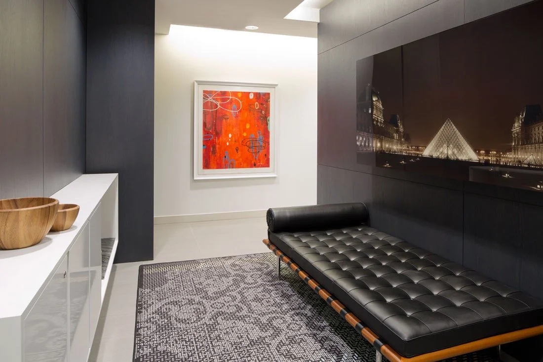 Modern interior with a black leather daybed, abstract artwork on a white wall, and a large black panel with cityscape reflections, with a white sideboard holding wooden bowls.