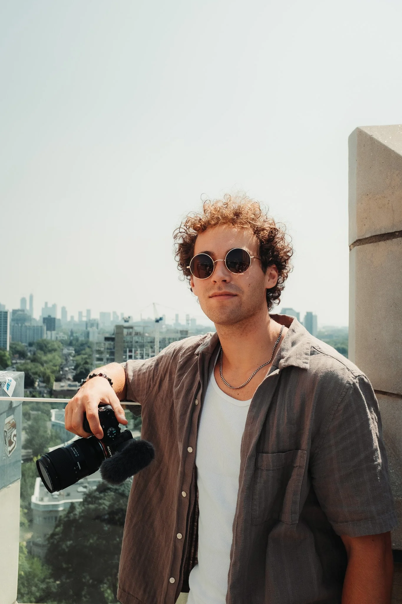 A young man with curly hair wearing sunglasses, a gray shirt, a white t-shirt, and jewelry, is standing outdoors on a rooftop with a city skyline in the background. He's holding a camera with a microphone attached.