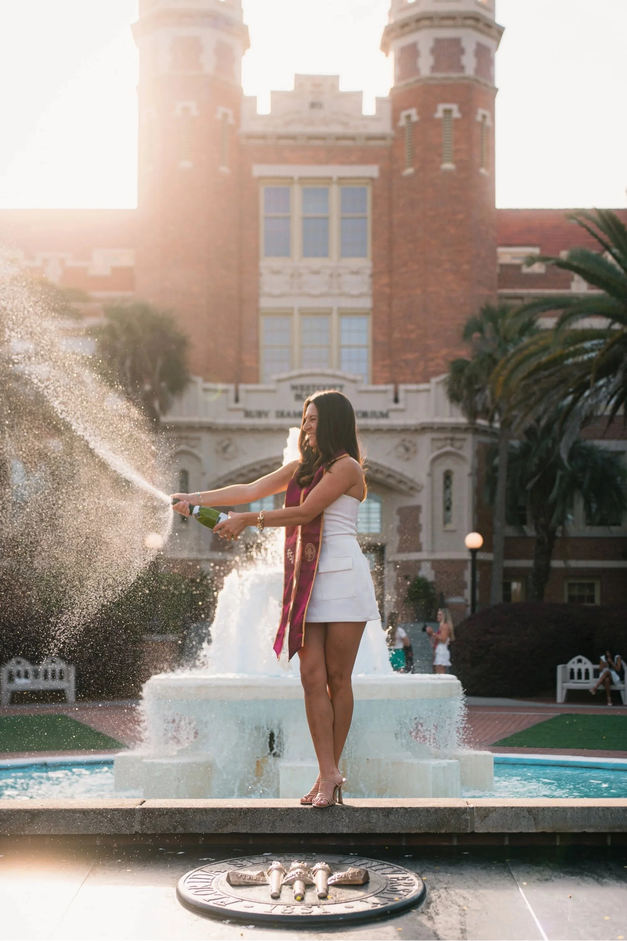 A woman in a white dress and sash celebrating at a graduation ceremony, standing on a fountain and opening a bottle of champagne, with a brick university building in the background during sunset.