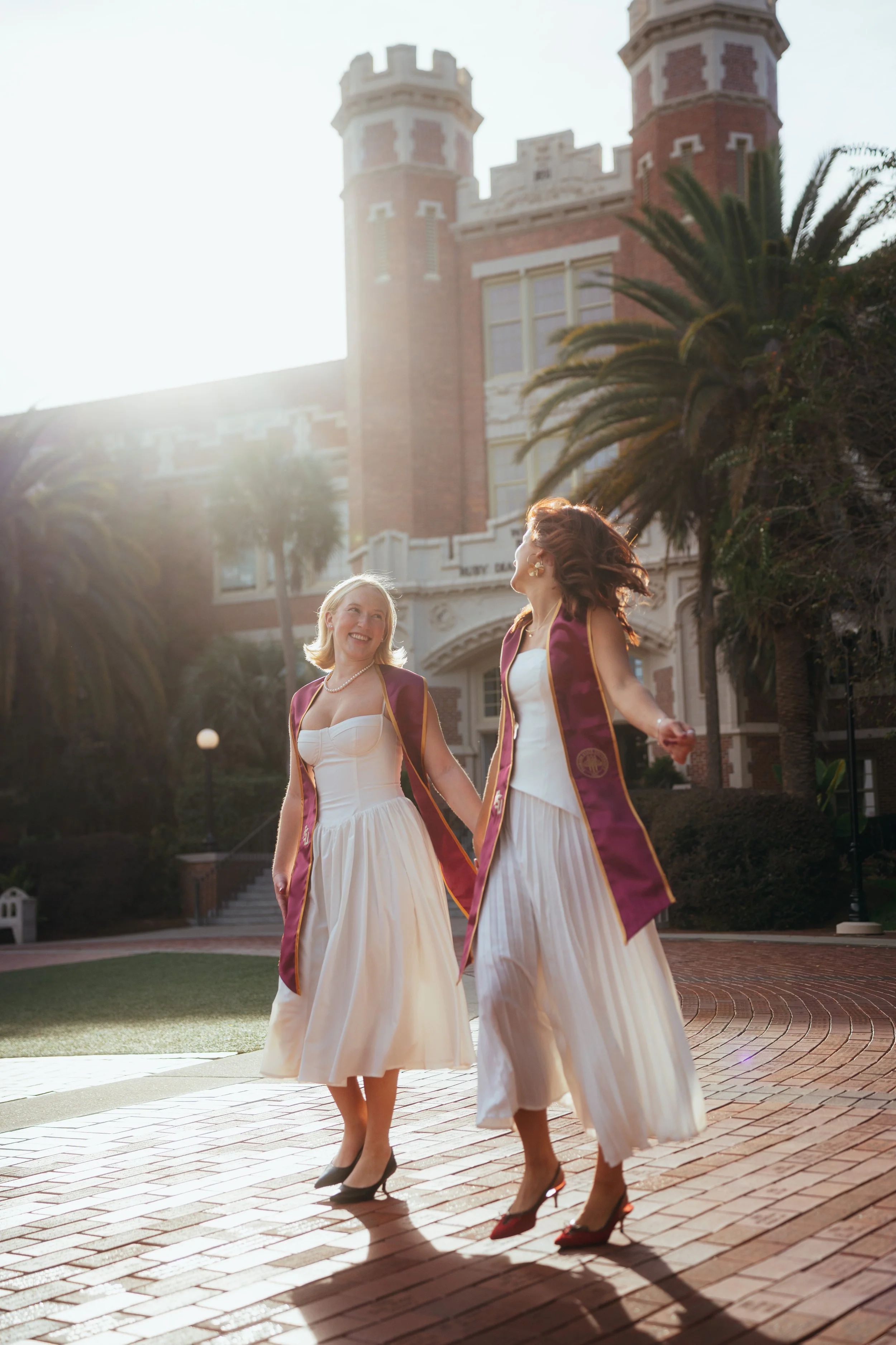 Two women in formal dresses and graduation stoles holding hands and dancing on a brick pathway in front of a historic building with towers and palm trees, with sunlight glowing behind them.