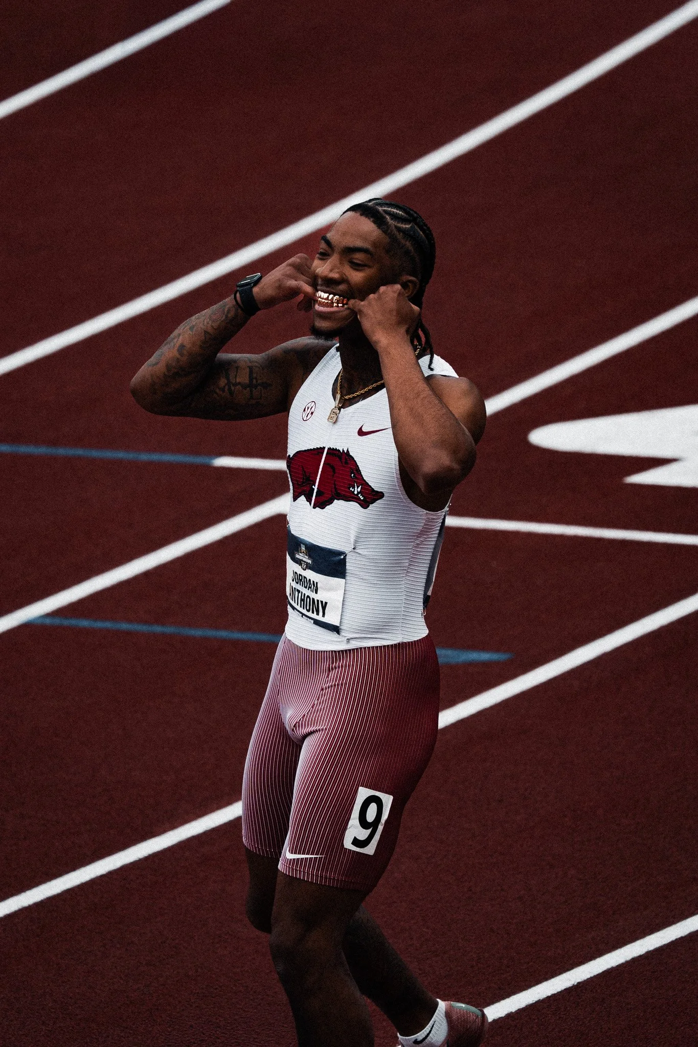 A male athlete smiling and adjusting his earbuds on a red running track, wearing a white and maroon Arkansas Razorbacks jersey with the number 9.