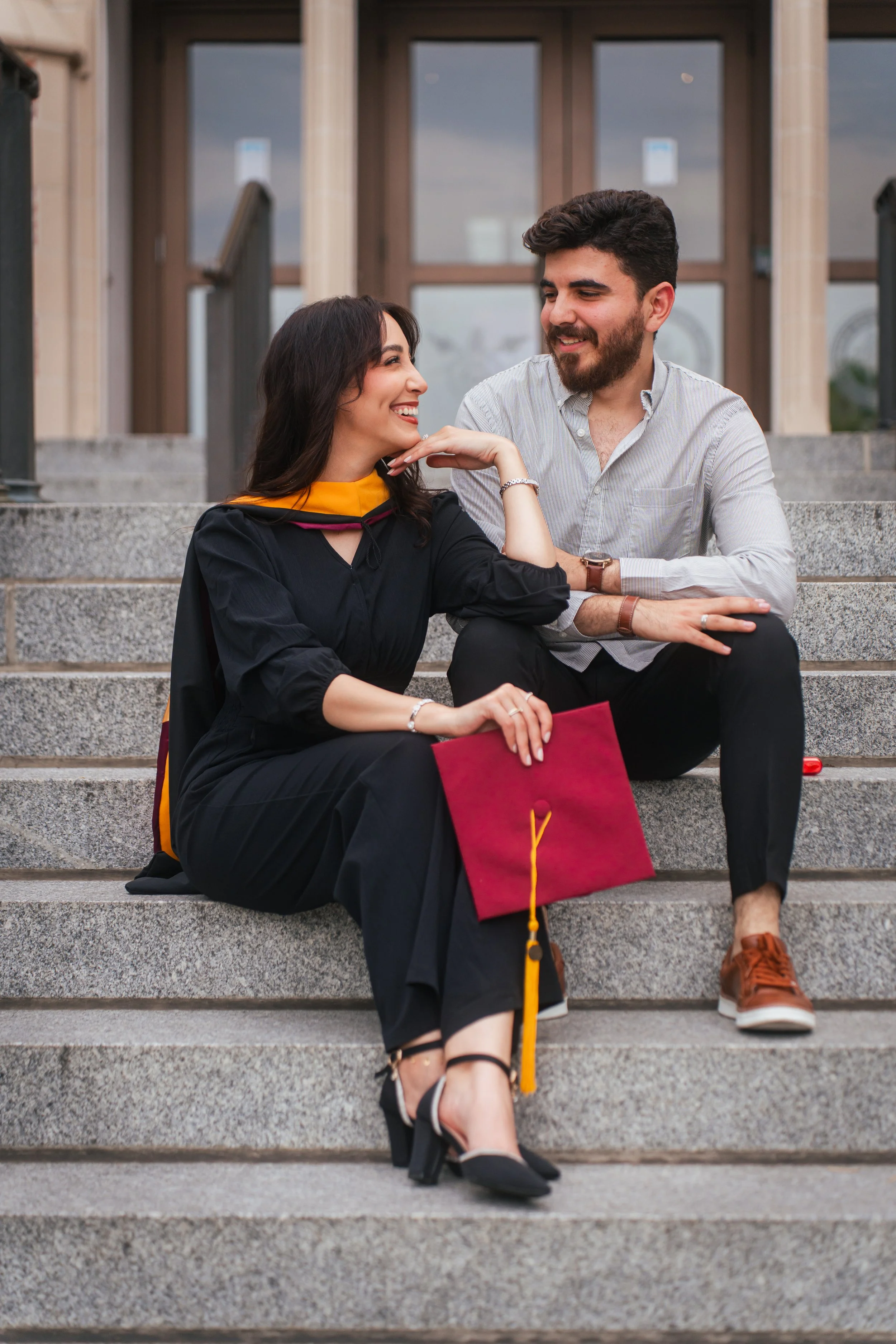 A woman in a graduation gown and cap sitting on stairs with a man in casual attire, holding a red diploma cover, celebrating her graduation.