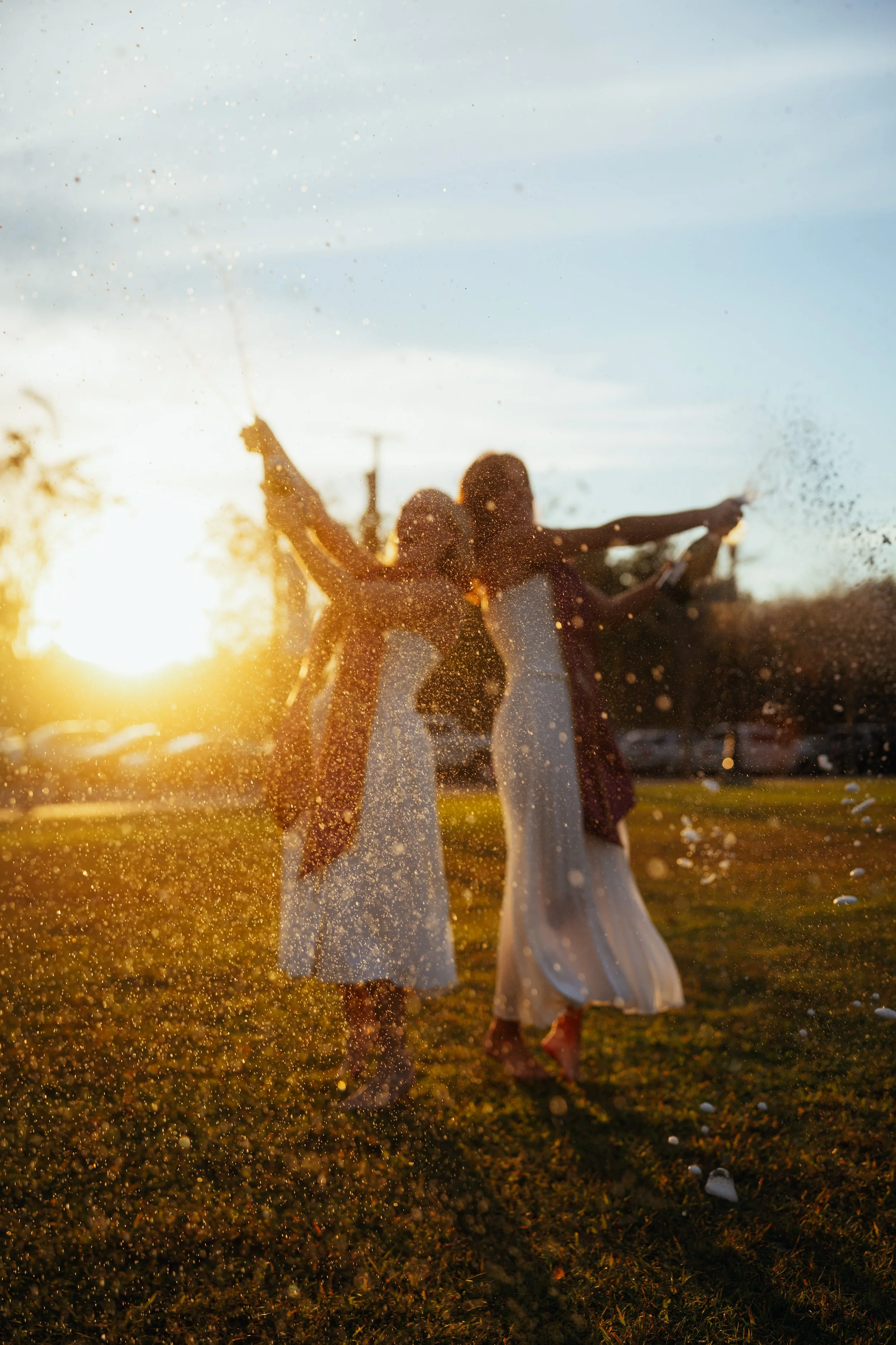 Two women in white dresses playing and splashing water on a grassy field during sunset.