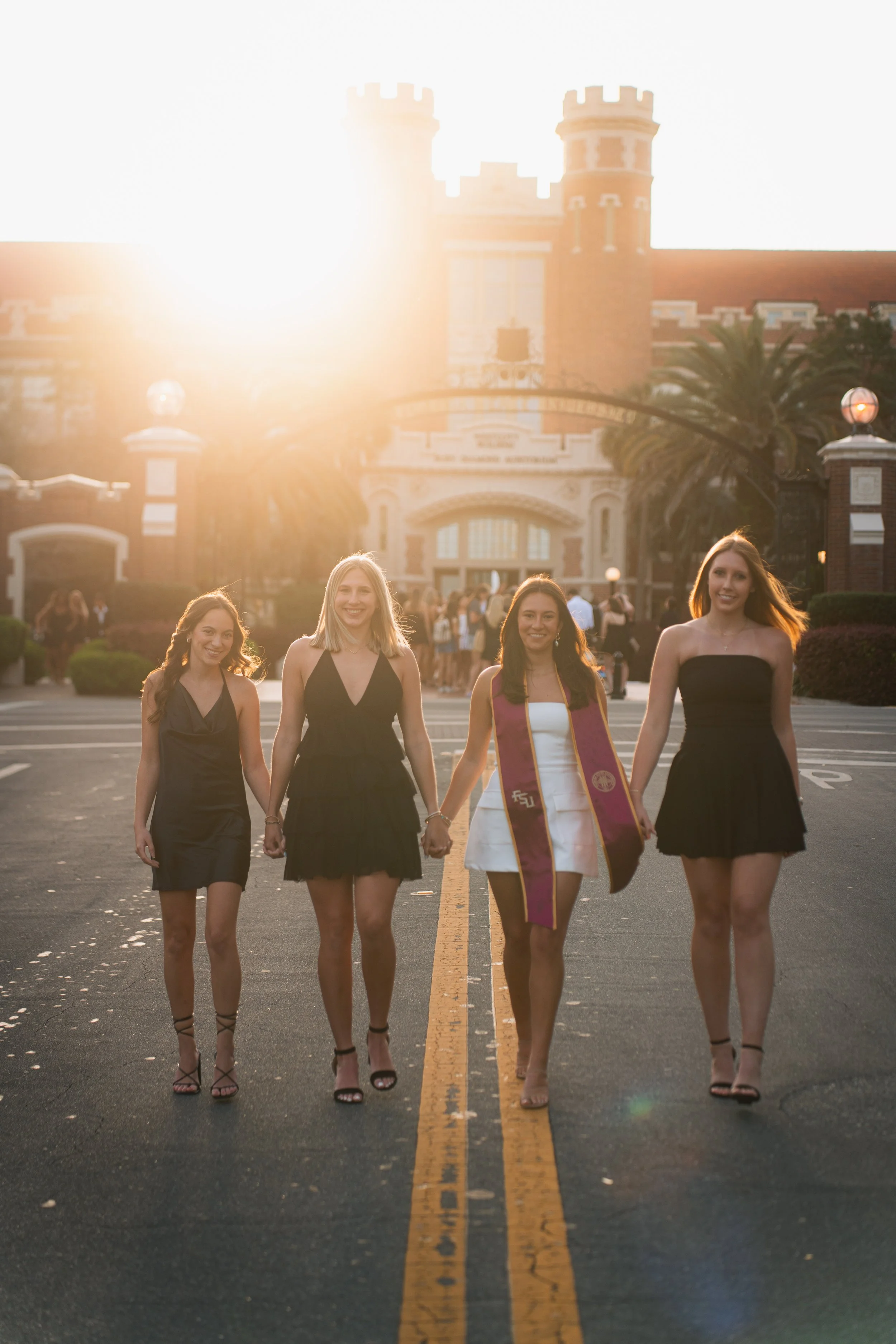 Four young women in evening dresses walking hand-in-hand on a campus street during sunset; one is wearing a graduation stole, and the background features a historic university building with turrets.