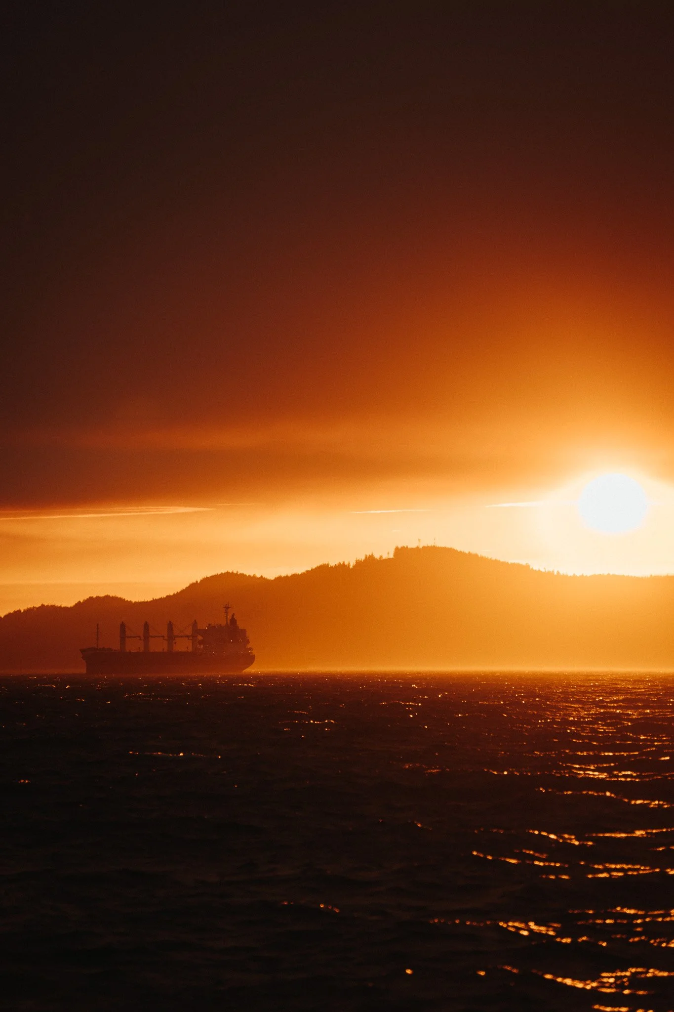 A ship sailing near a mountainous coastline during sunset with an orange and yellow sky.
