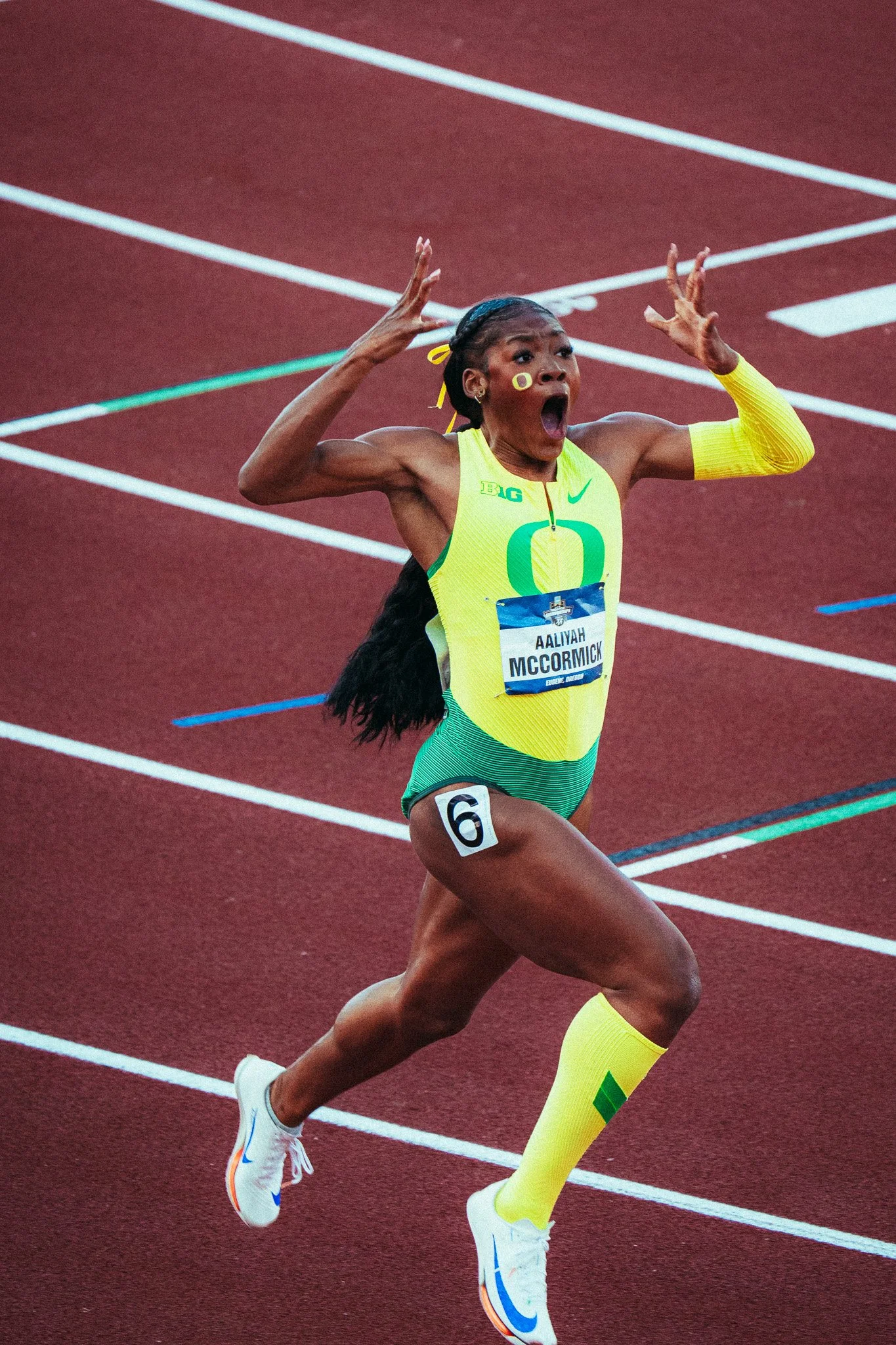 A female track athlete with long hair in a yellow and green uniform, numbered '6', running on a red track with her mouth open and arms raised in an exclamation.