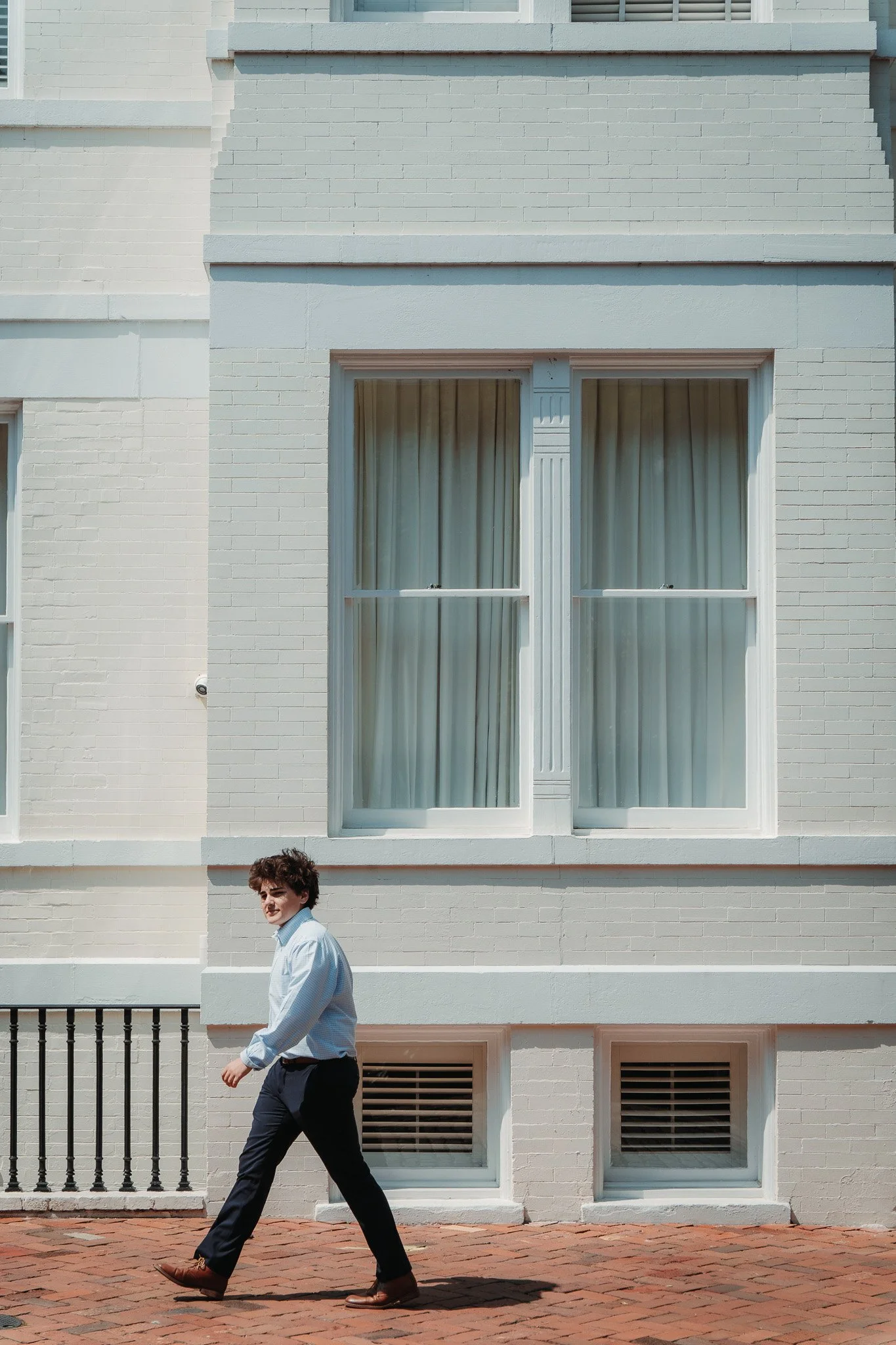 A man in a light blue shirt and dark pants walking past a light-colored building with large windows and brick sidewalk.