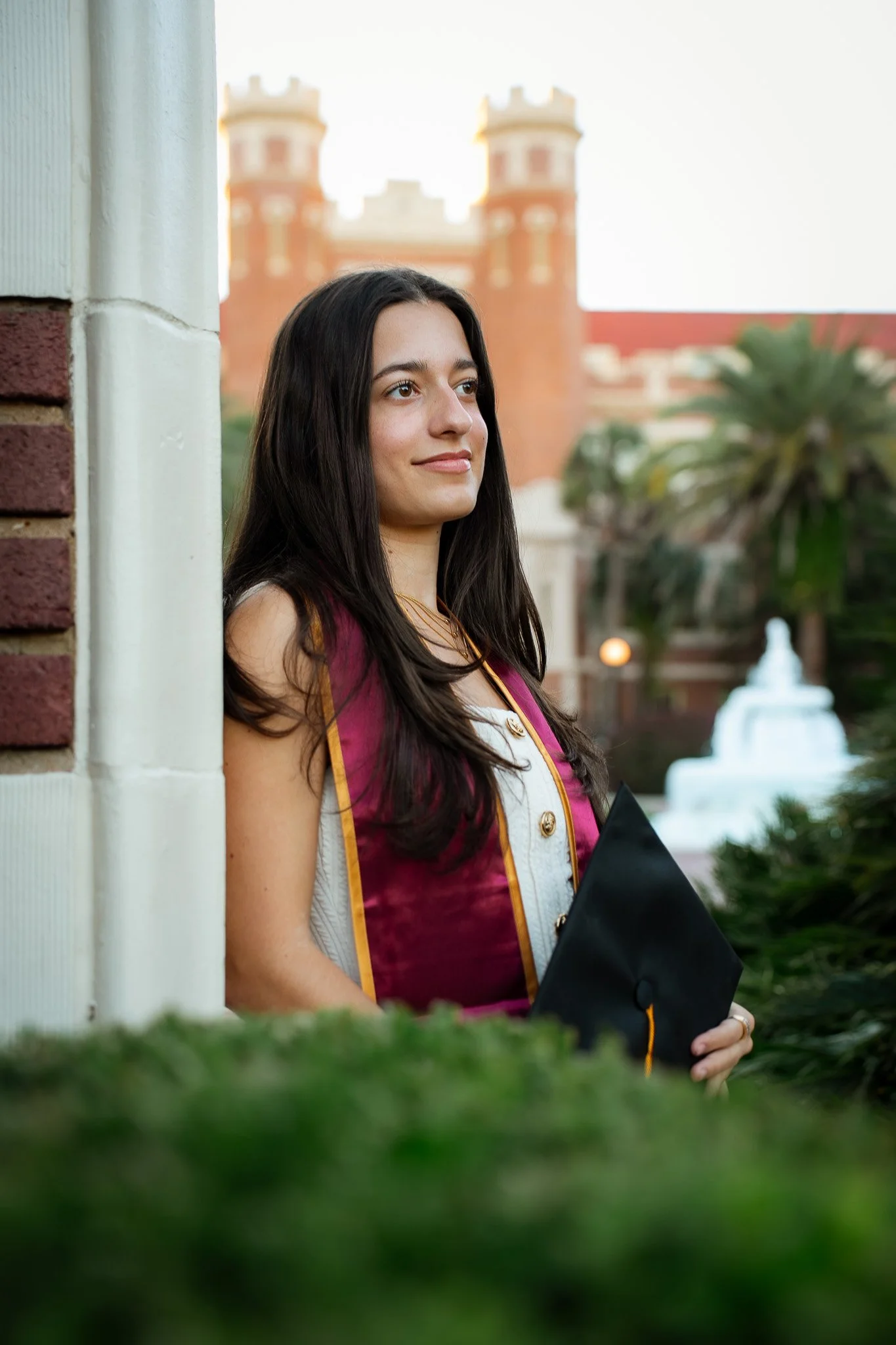 Young woman in a graduation gown holding a diploma, standing outside in front of a castle-like building.