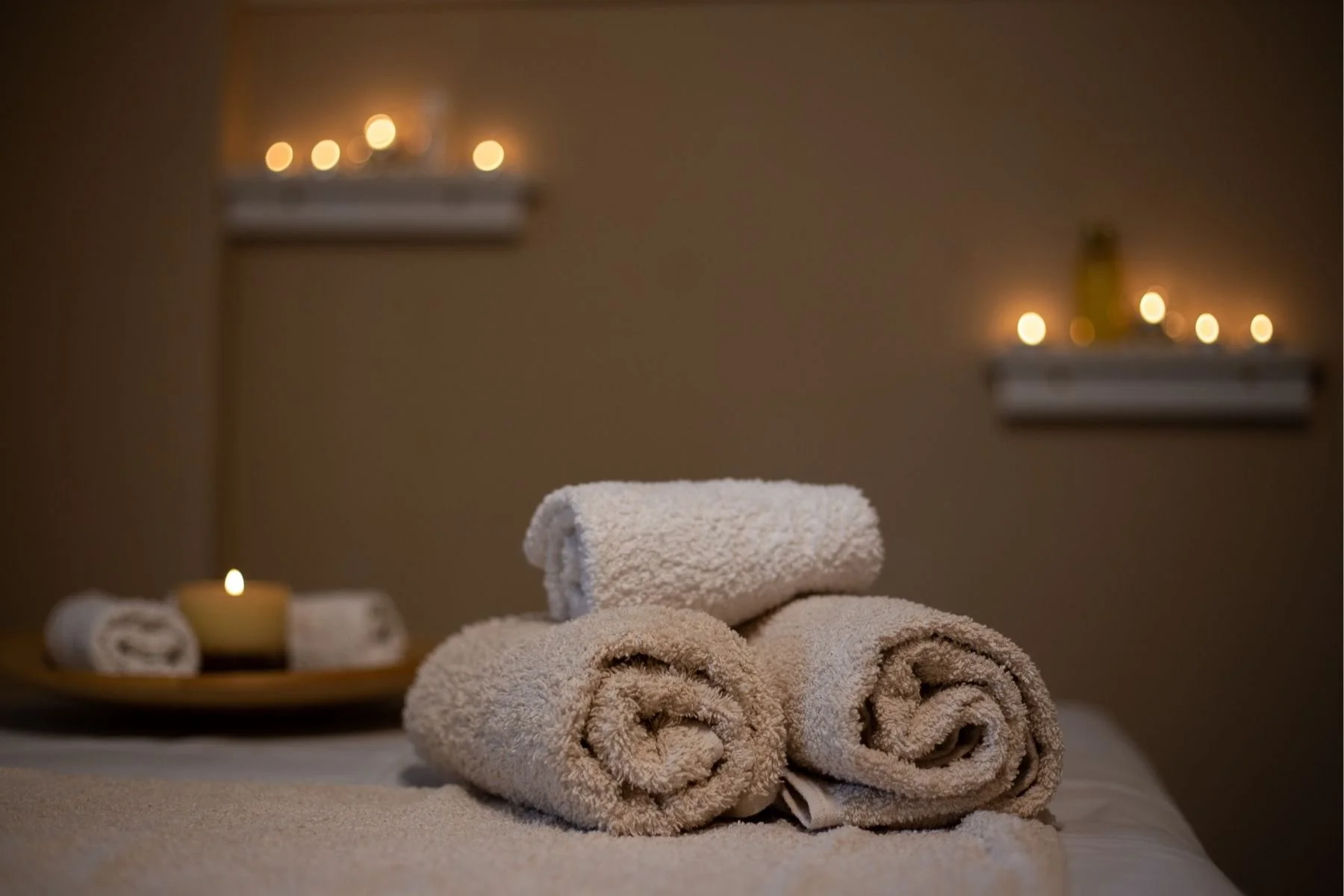 Three rolled towels, two beige and one white, placed on a bed in a dimly lit room with candles on a tray and shelves in the background, creating a calming spa-like ambiance.