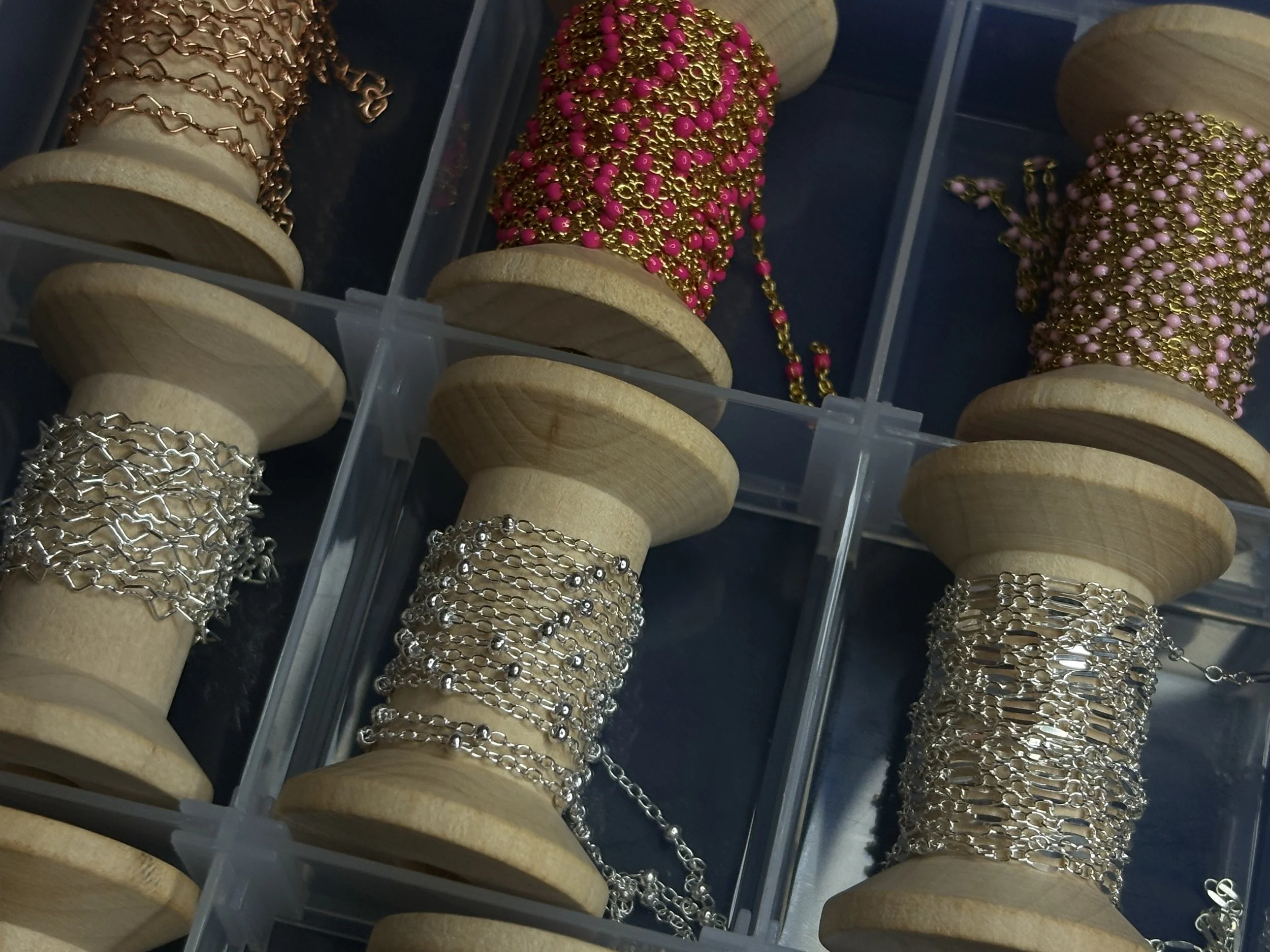 Multiple spools of jewelry chains and necklaces in silver, gold, and pink colors displayed in a transparent plastic container.