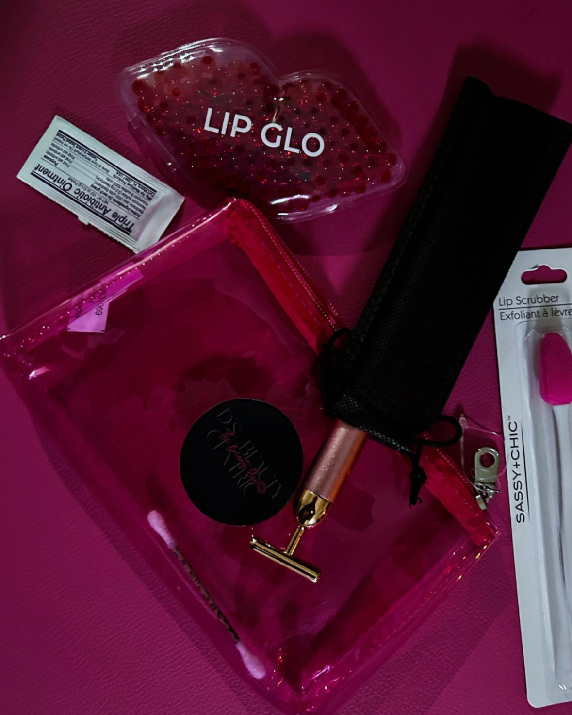 Collection of pink and black beauty items on a pink surface, including a heart-shaped lip gloss container labeled 'LIP GLO,' a pink lip roller, a black zippered pouch, a lip scrubber in packaging labeled 'SASSY4CHIC,' and a small packet of lip additi