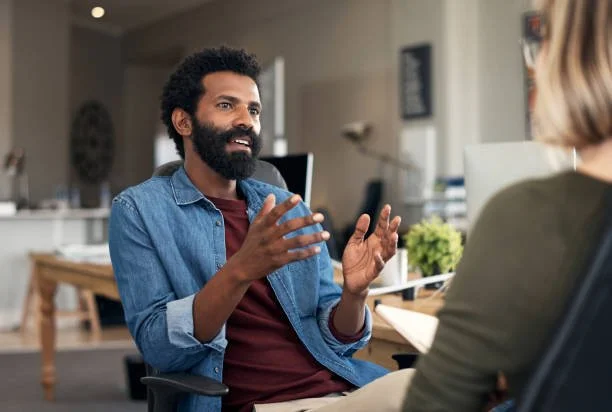 Man with curly hair and beard speaking in a conversation or interview with a woman in an office setting.