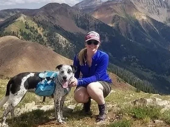 A woman in outdoor clothing and sunglasses kneeling next to a black and white dog with a blue backpack on a mountain trail, with mountains and a valley in the background.