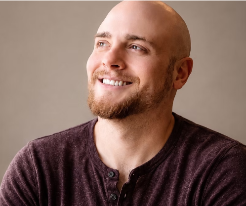 Close-up of a smiling, bald man with a beard wearing a dark casual shirt, looking slightly to the right against a neutral background.