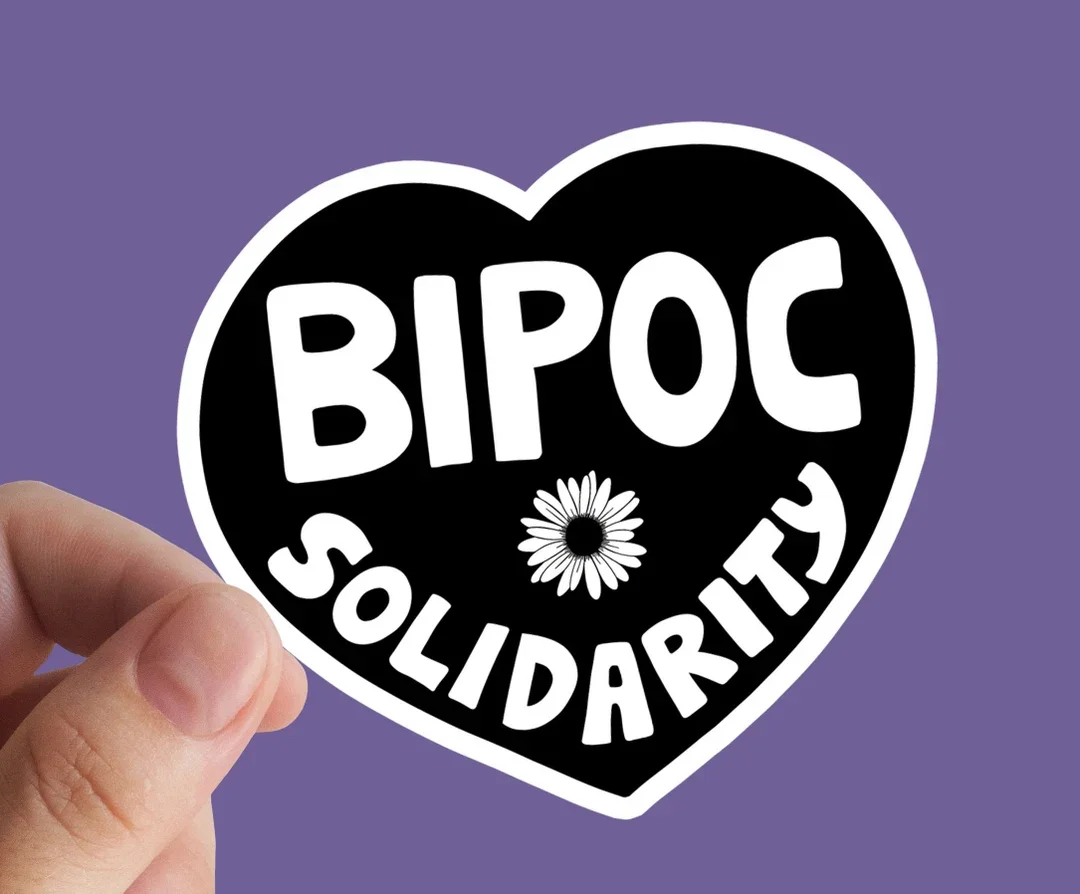A person holding a heart-shaped sticker with the words "BIPOC SOLIDARITY" and a daisy flower in the center, against a purple background.