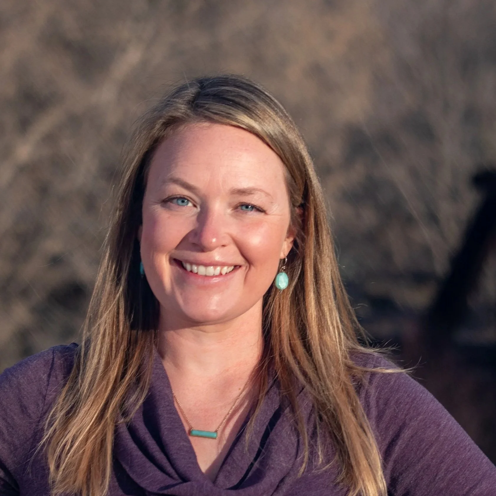 Close-up of a smiling woman with long, light brown hair and light blue eyes, wearing turquoise earrings, a turquoise necklace, and a purple top, outdoors with a natural, blurry background.