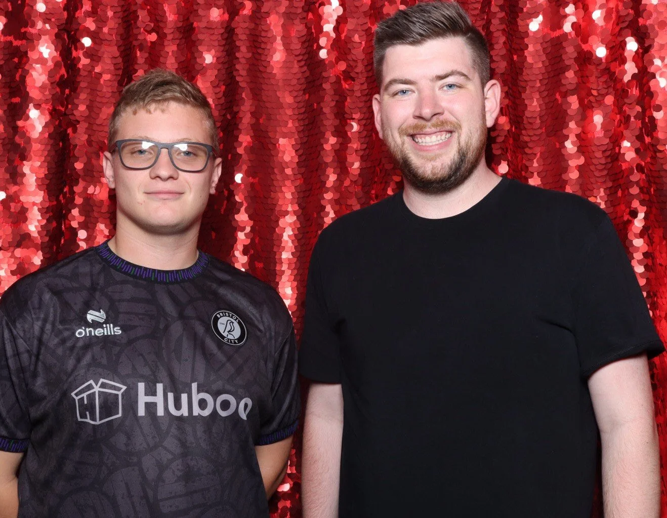 Two men standing side by side against a red sequin backdrop, one wearing glasses and a black sports jersey, the other smiling in a black t-shirt.