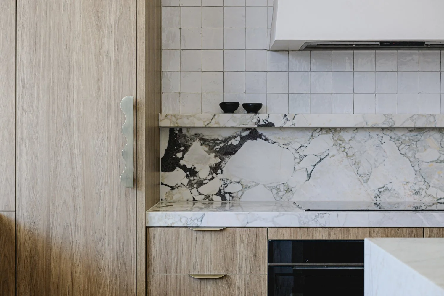 Close-up of a modern kitchen with wood cabinets, marble countertops and backsplash, and two small black bowls on the counter.