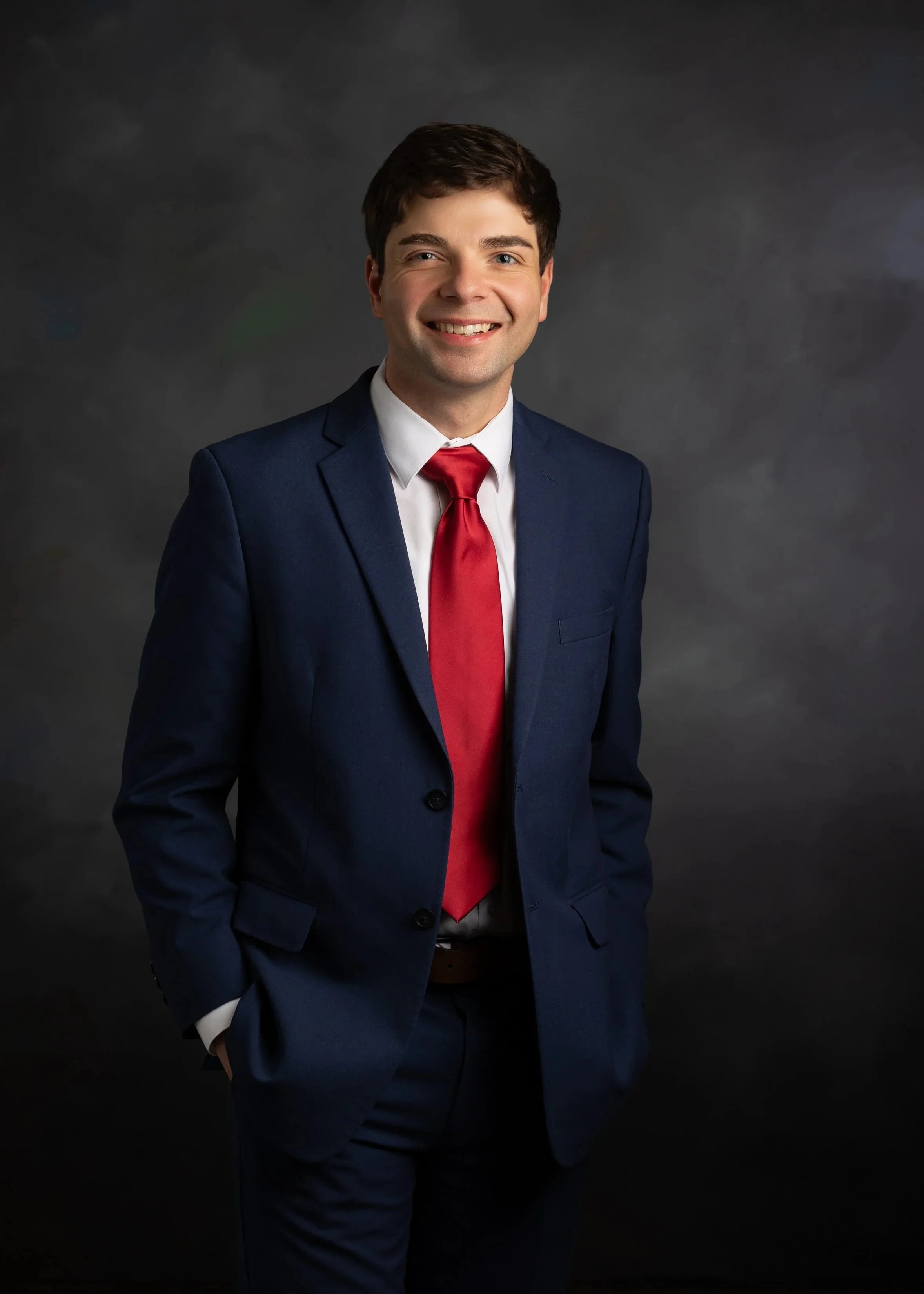 Young man in a navy suit, white shirt, and red tie standing against a dark background, smiling and posing for a professional headshot.