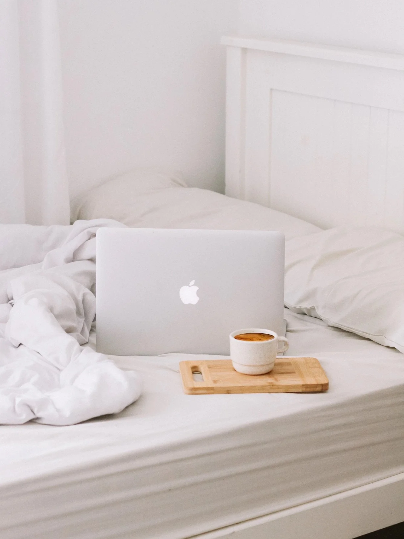 A cozy bed with white sheets, a pillow, an open MacBook, a cup of coffee on a wooden tray, and a white headboard in a bright room.