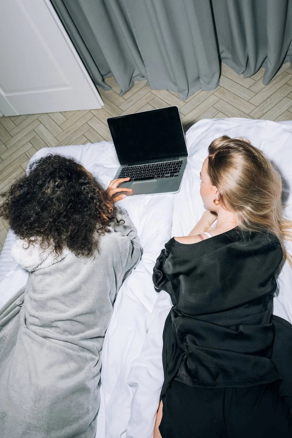 Two women lying on a bed, looking at a laptop computer together.
