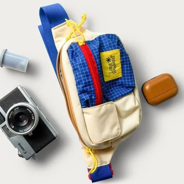 A small beige and blue backpack with a yellow zipper and a yellow patch labeled "Lodge Weekend" is surrounded by a vintage camera, a white flash, and a brown case on a white background.
