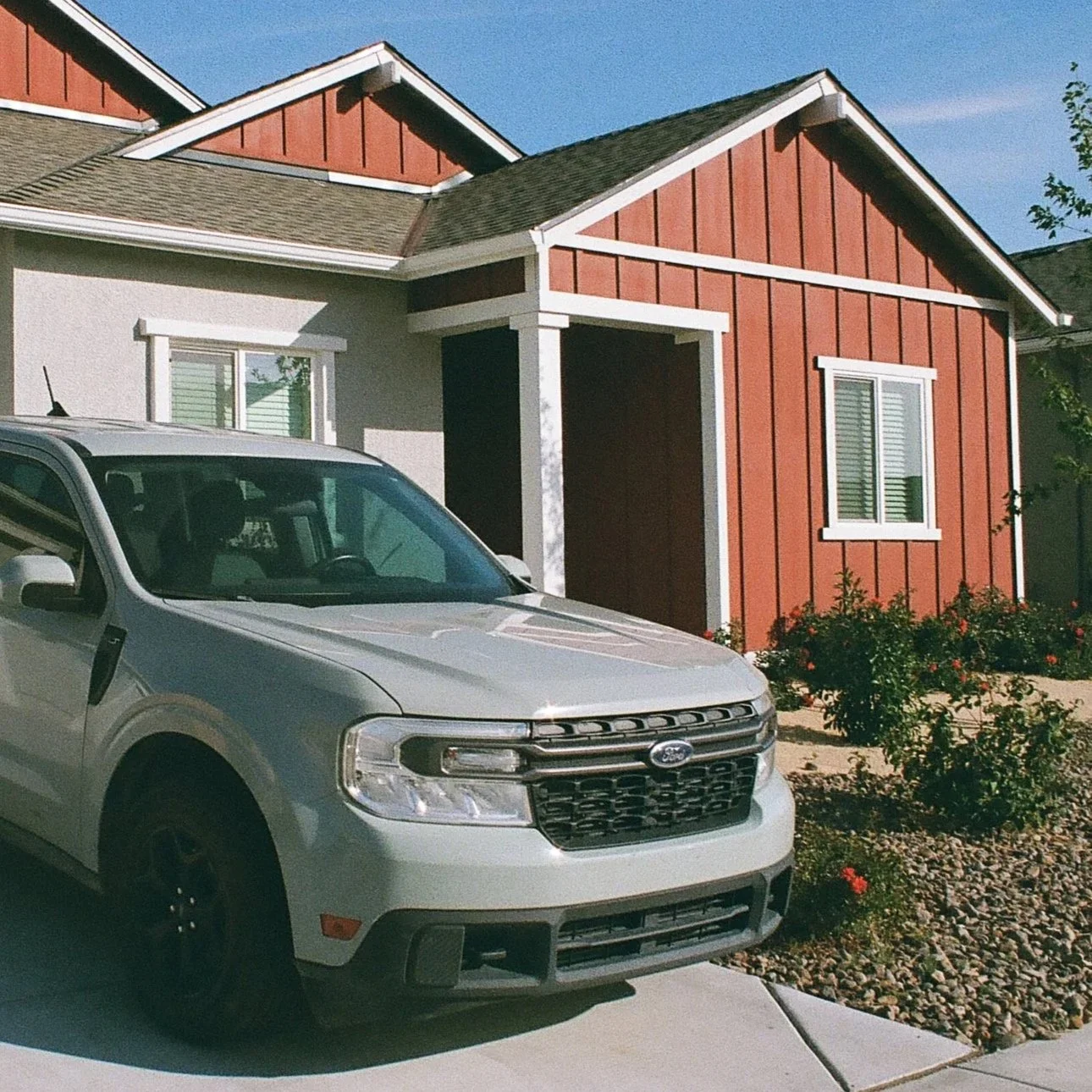 A white SUV parked in front of a house with red and white exterior walls, two windows with shutters, and landscaped bushes with flowers.