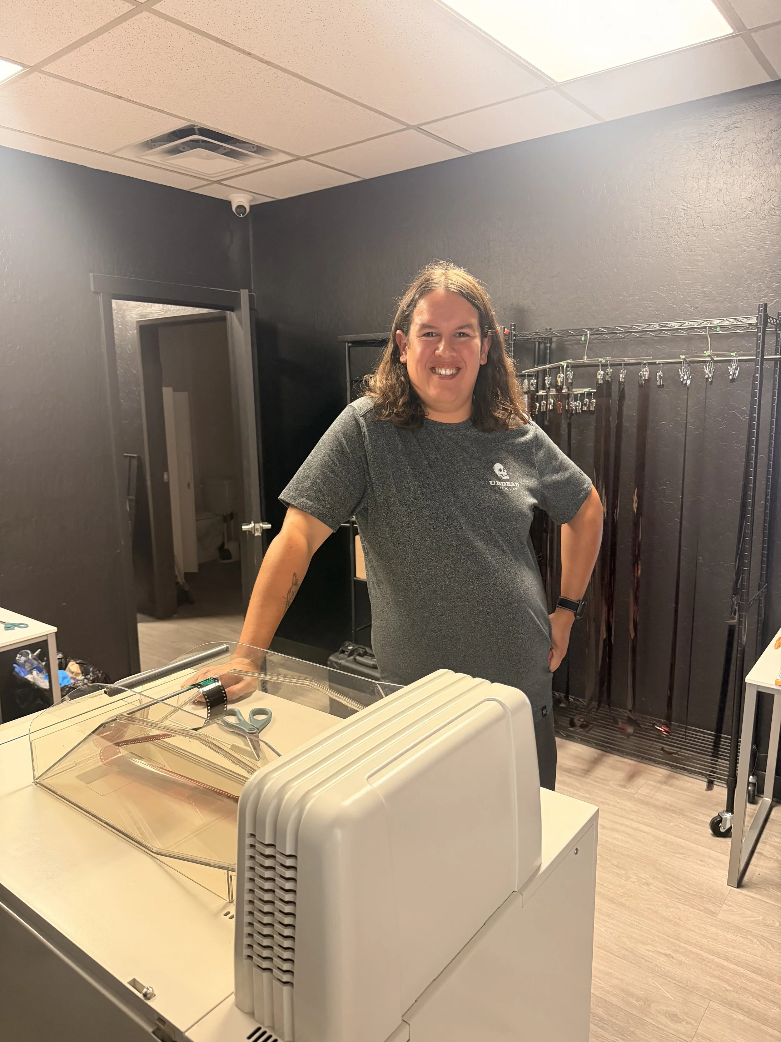 Smiling person with long hair standing inside a clothing or accessory store. There are racks with belts behind them and a display with jewelry. The person is wearing a gray T-shirt with a skull and text logo.