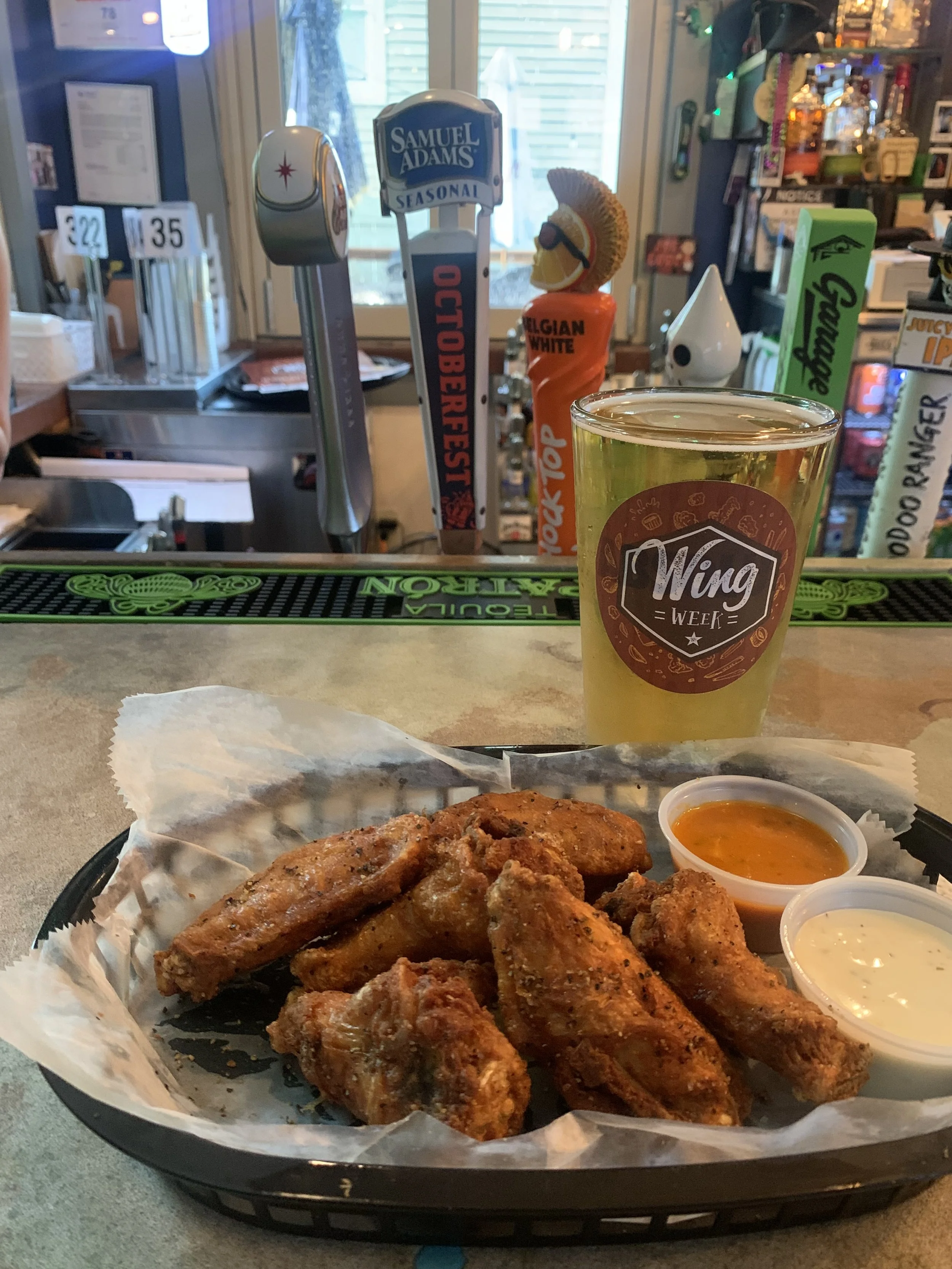 Basket of fried chicken wings with two dipping sauces, a glass of beer, and bar decor in the background.