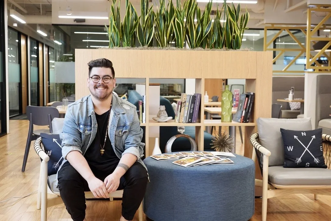 A man sitting on a chair in a modern office space, smiling at the camera. Behind him is a wooden bookshelf with plants on top, decorative items, and books. The office has glass partitions, wooden floors, and contemporary furniture.