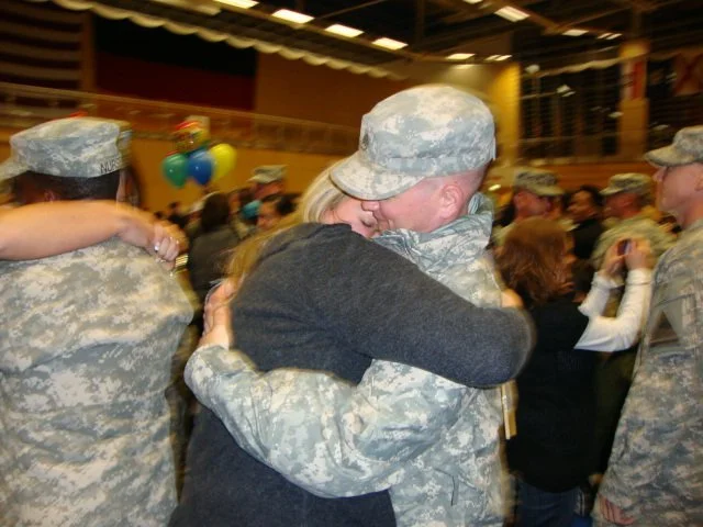 Military personnel in uniform hugging and holding a woman in celebration at a gathering.