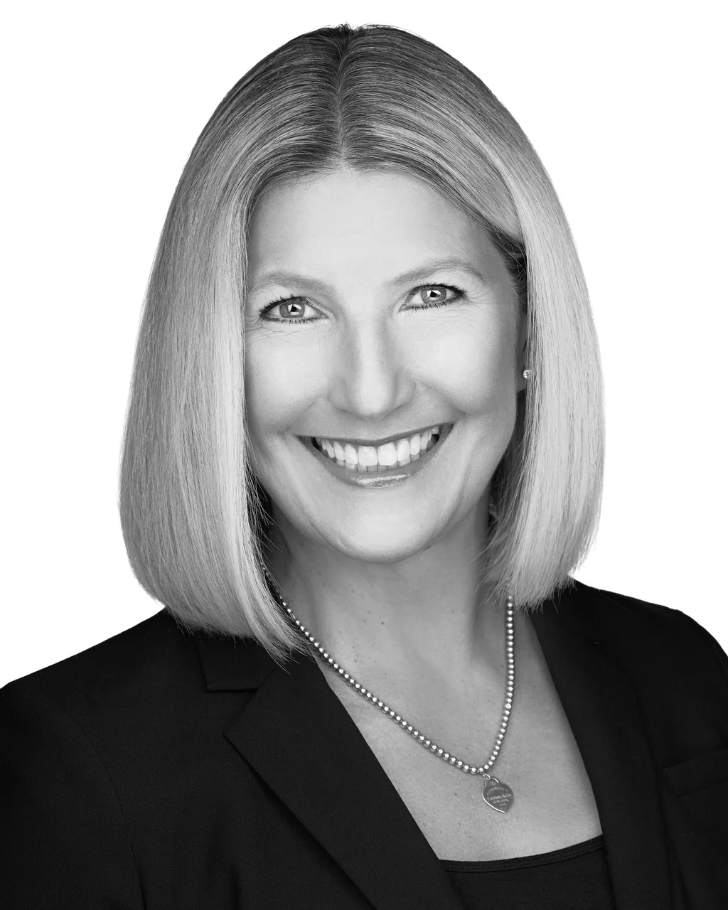 Black and white headshot of a woman with shoulder-length straight hair, smiling, wearing a dark blazer and a beaded necklace with a heart-shaped pendant.